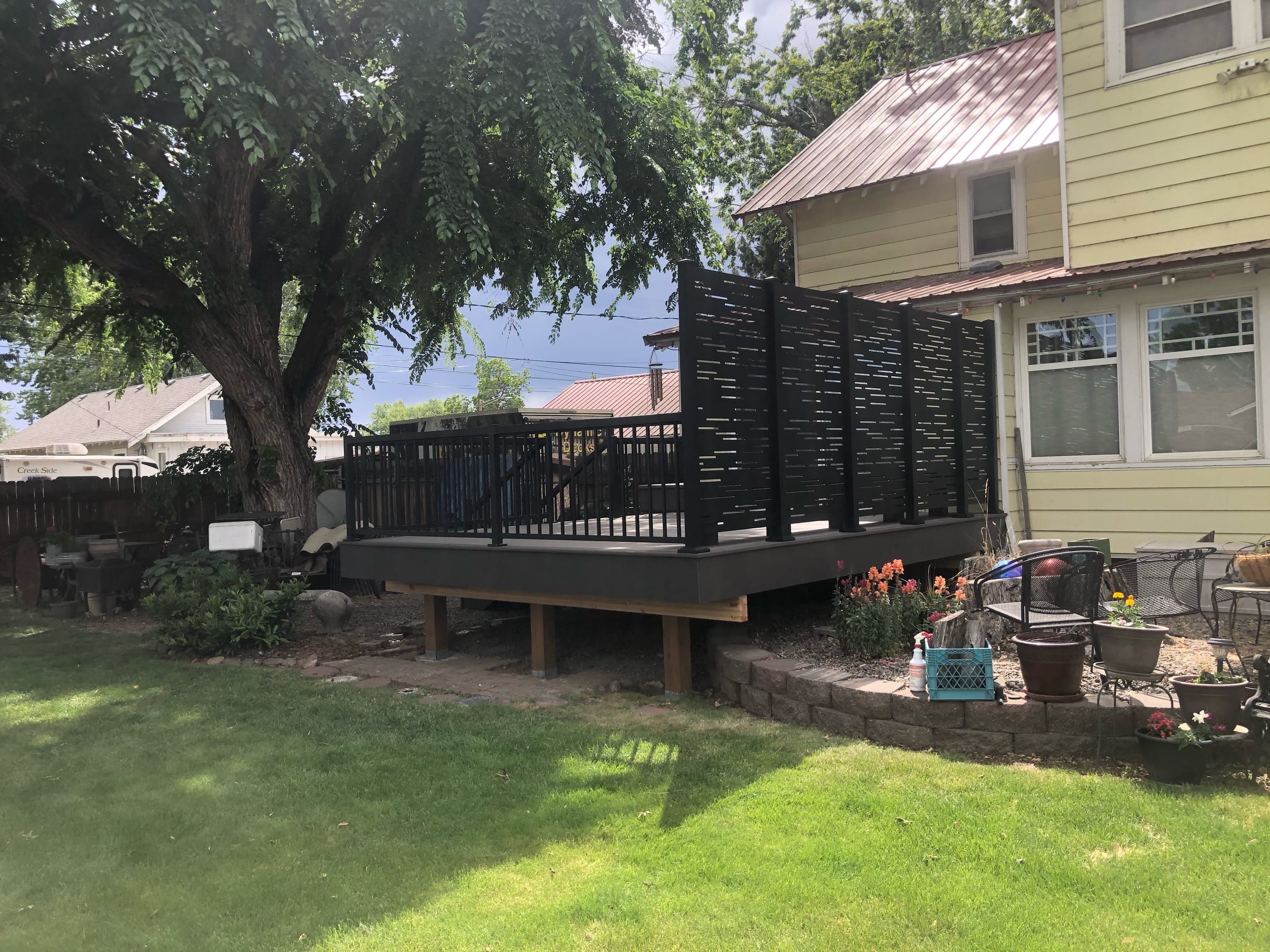 Raised gray composite deck with black picket railing and tall black privacy screen beside yellow house and lawn