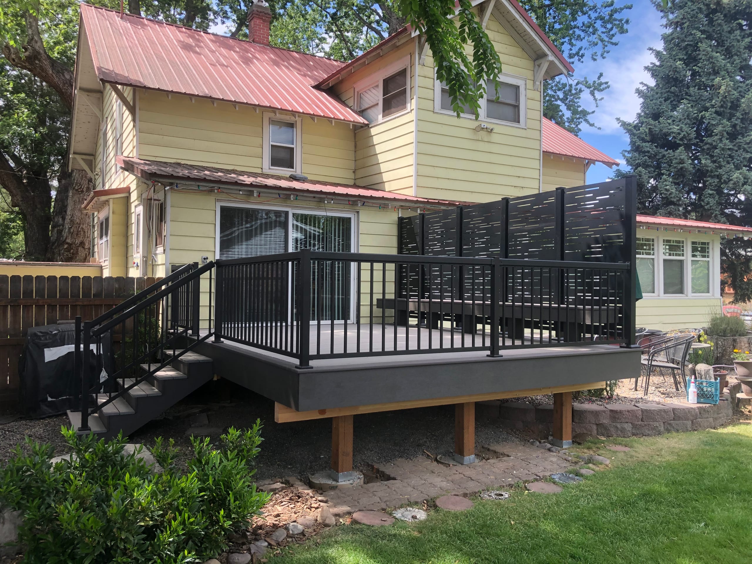 Raised gray deck with black aluminum railing and horizontal slat privacy wall behind a yellow house