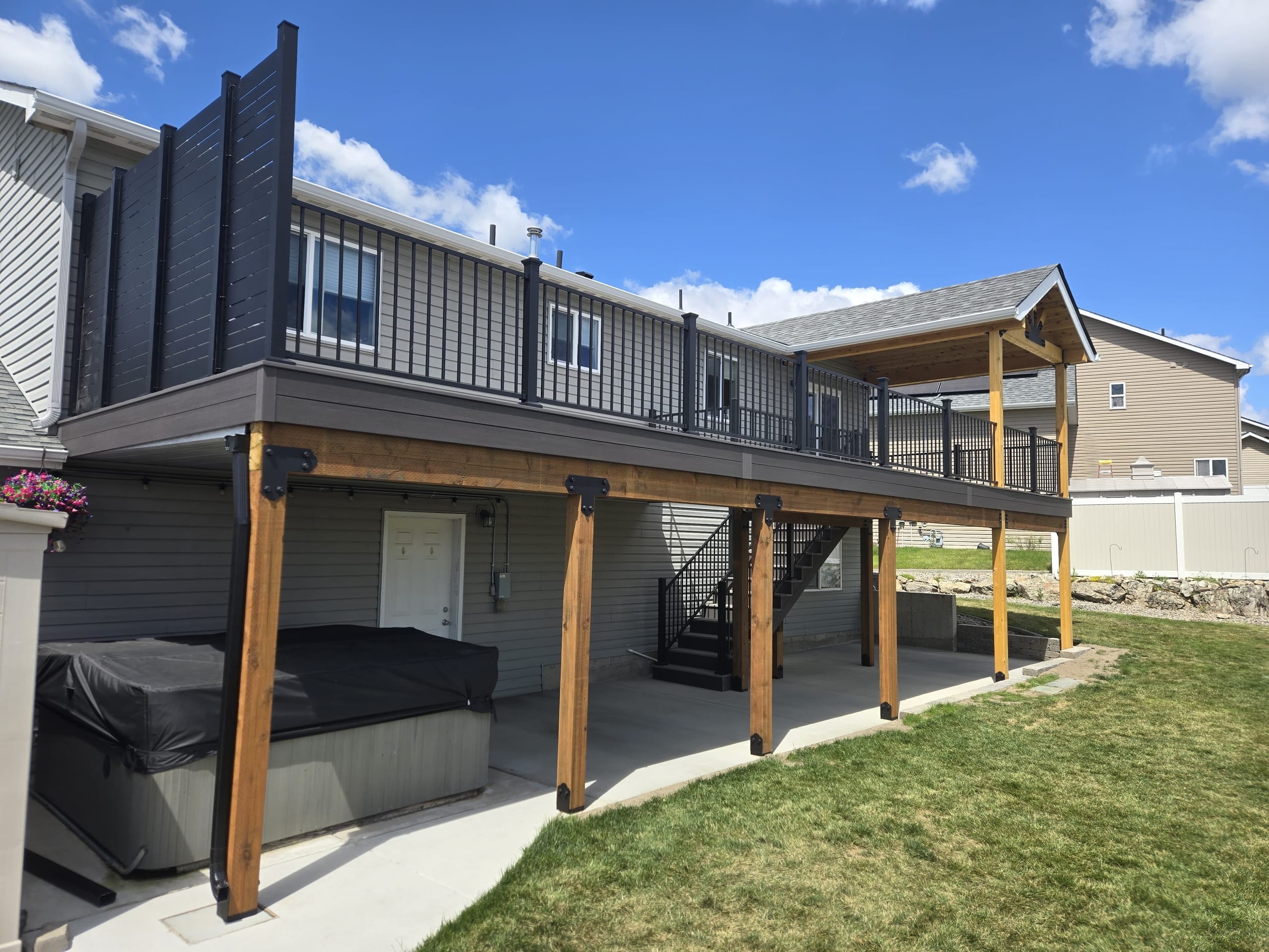 Raised gray deck on cedar posts with black railing, privacy screen, stairs and covered hot tub on patio beneath