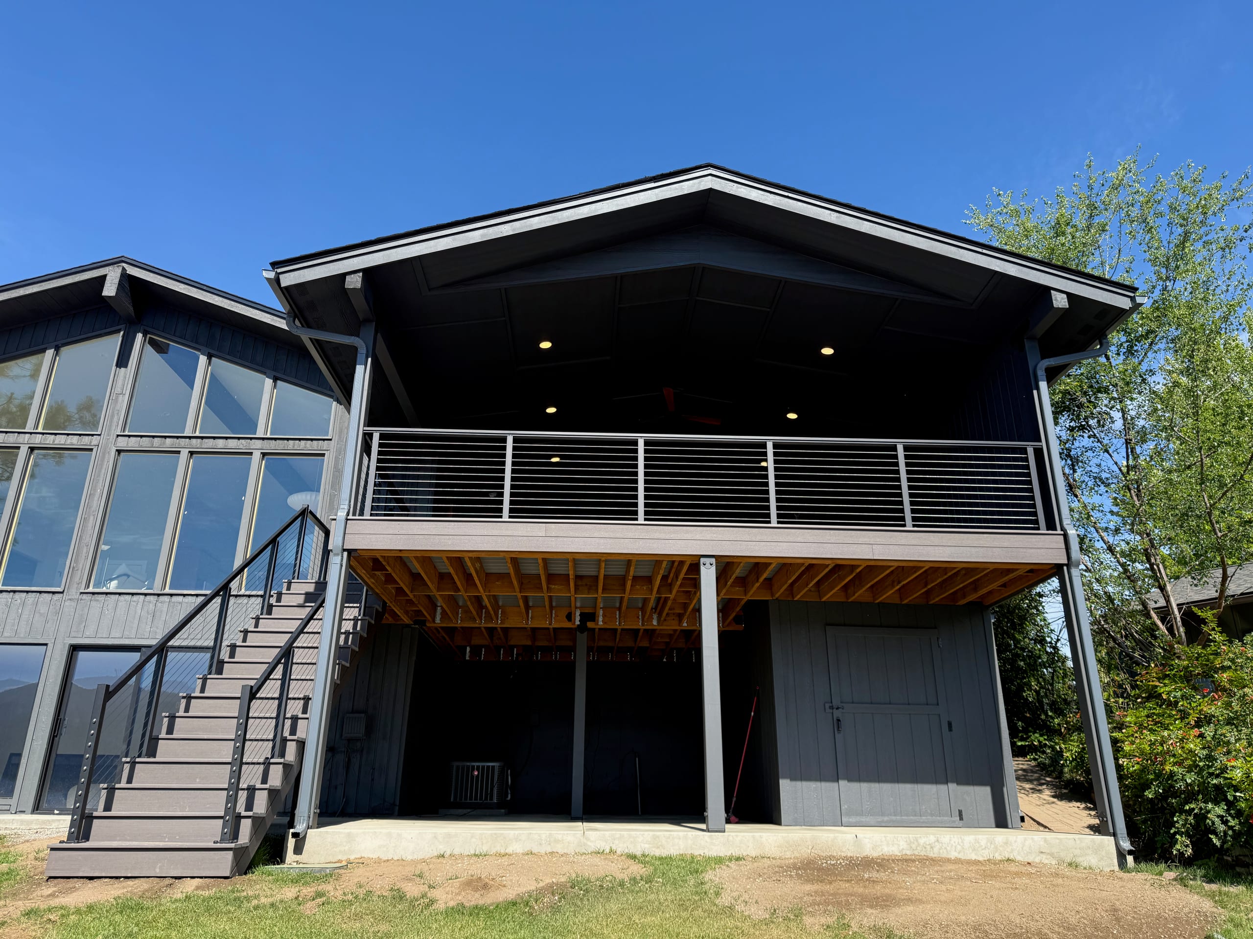 Rear elevation of dark house with gabled covered deck, cable railing, stairs and exposed underdeck, bright blue sky