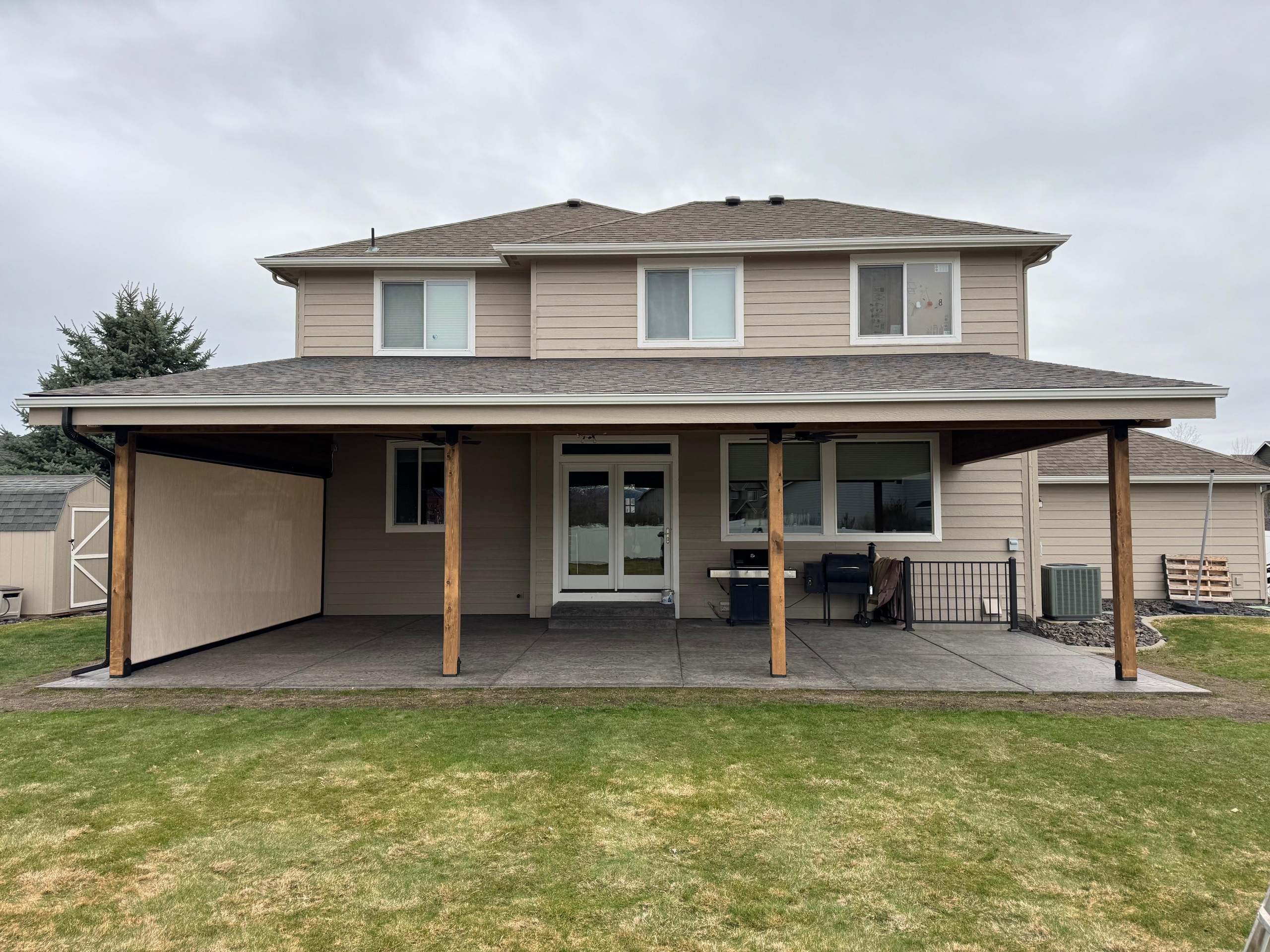 Rear view of two-story tan house with long low patio cover on timber posts, side sun shades and stamped patio, overcast