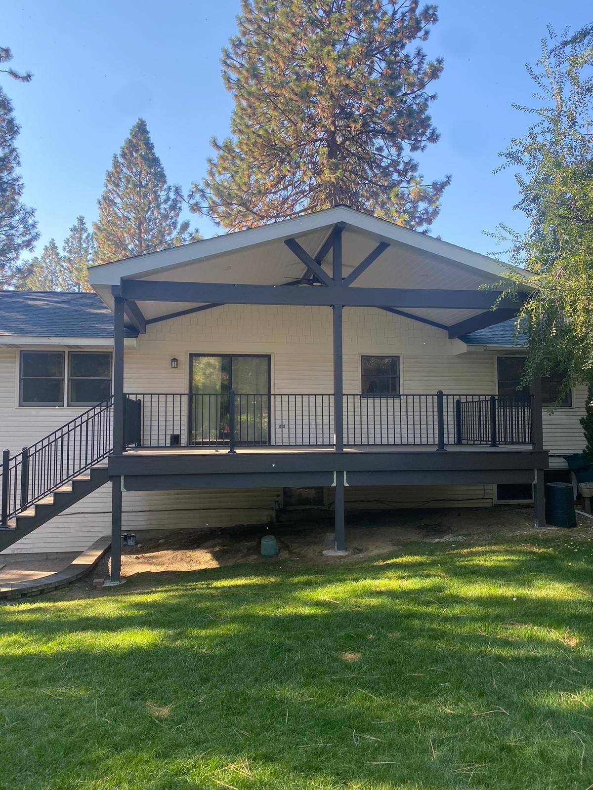 Rear of white house with gabled covered deck, dark trusses and black railing, green lawn and pine trees
