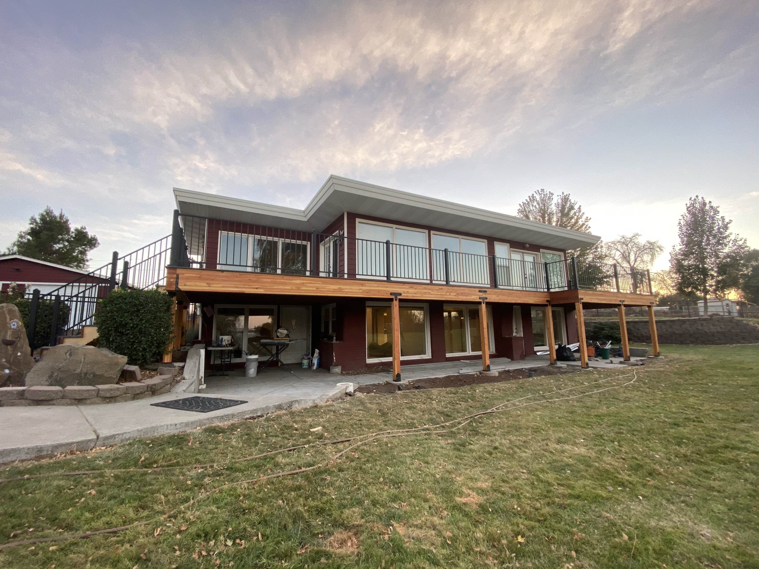 Red house with long elevated deck on cedar posts and covered patio below, lawn in foreground, dusk sky
