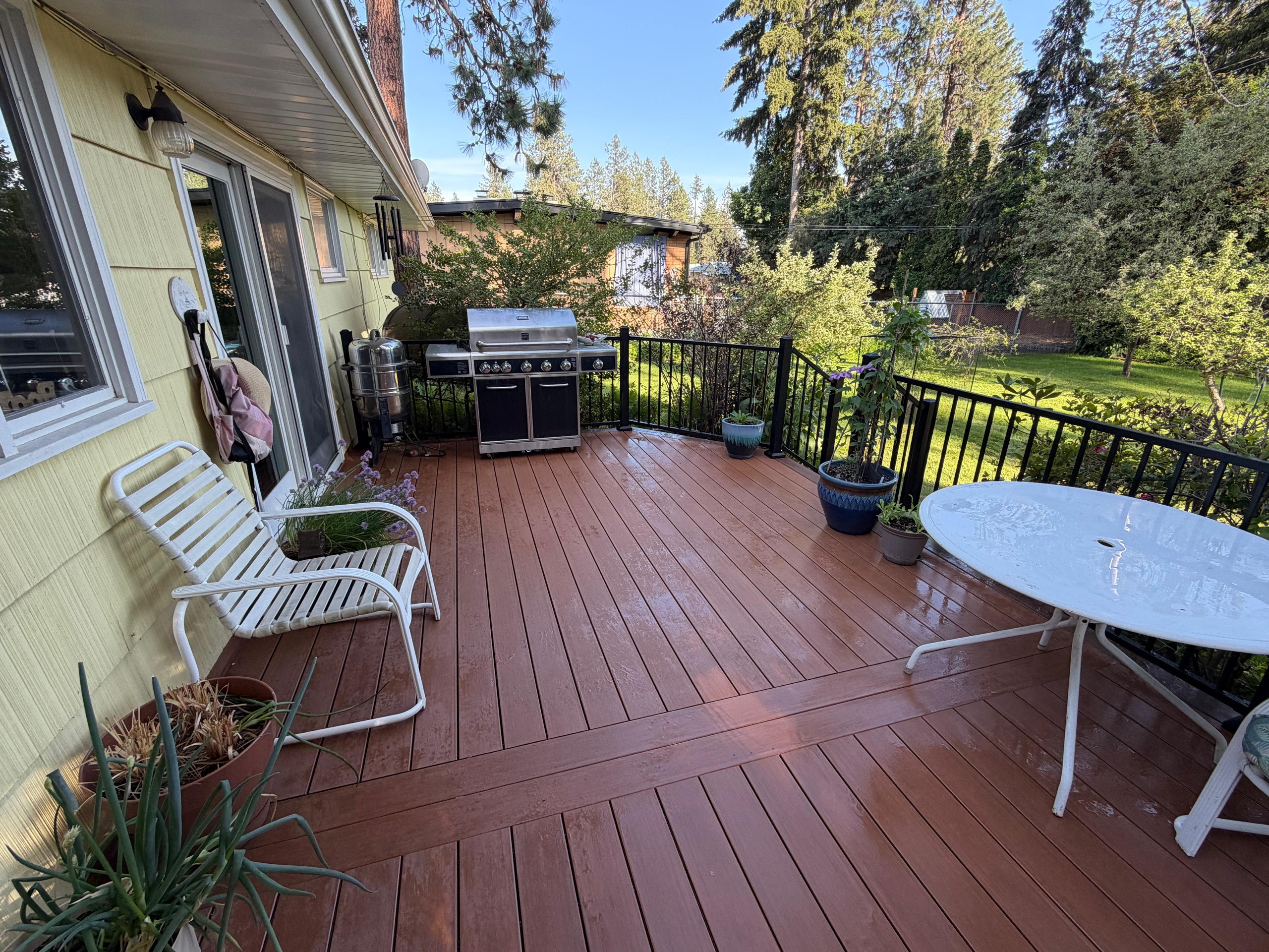Reddish-brown composite deck with black metal railing, patio table, chairs and grill at a yellow house, evening sun