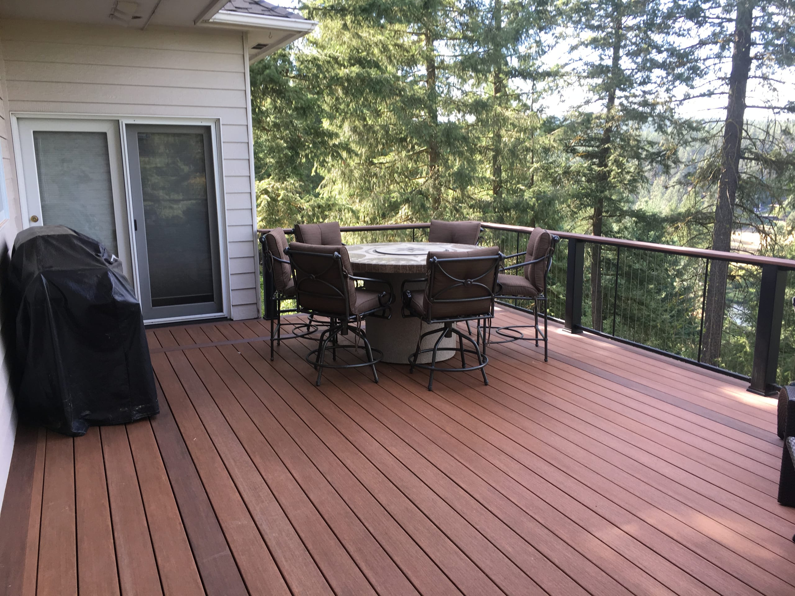 Reddish-brown composite deck with cable railing, patio dining table and tree-lined view from a white house