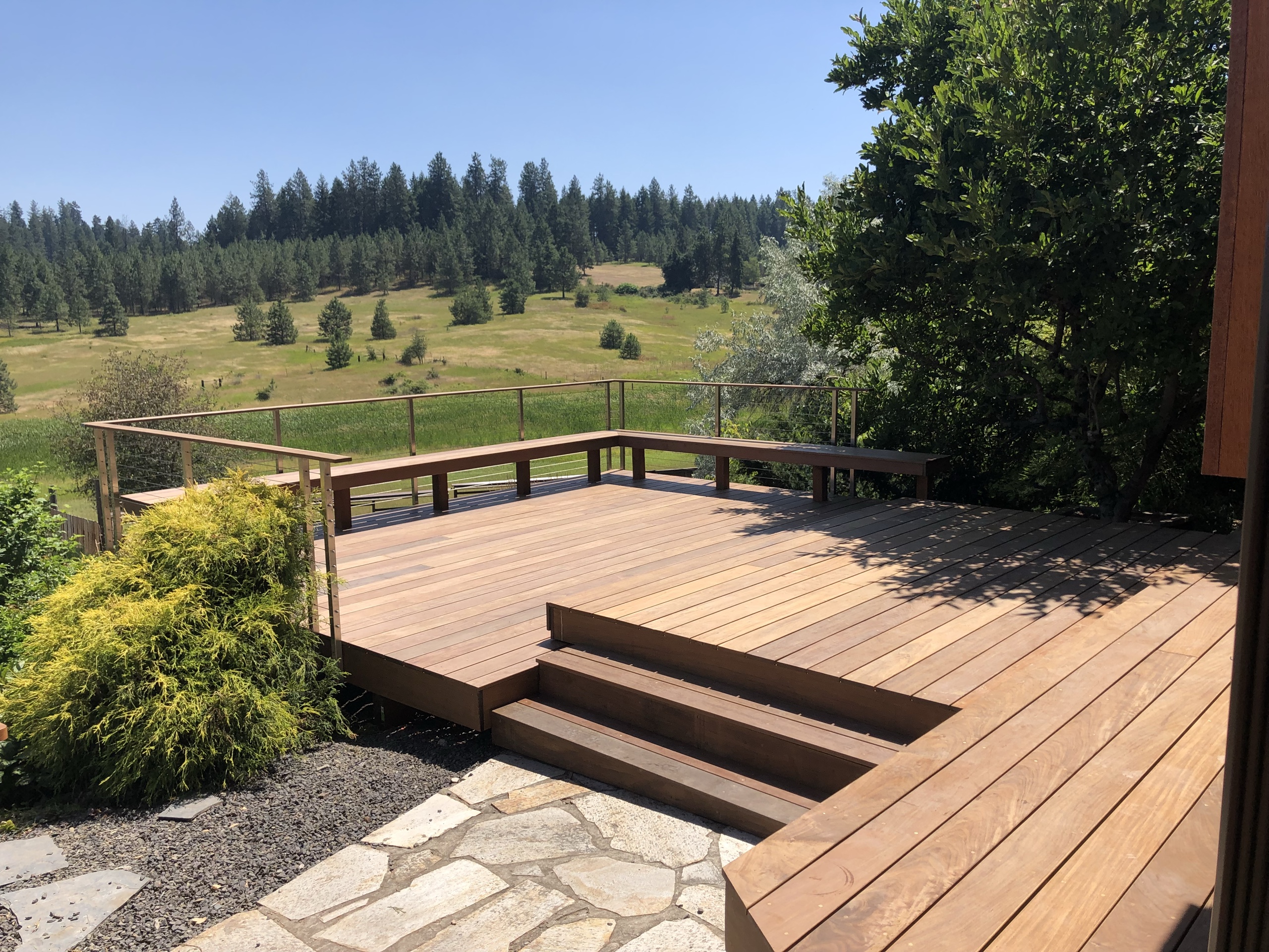 Reddish composite deck with wide steps and cable-style railing overlooking pasture, meadow and pines with flagstone path
