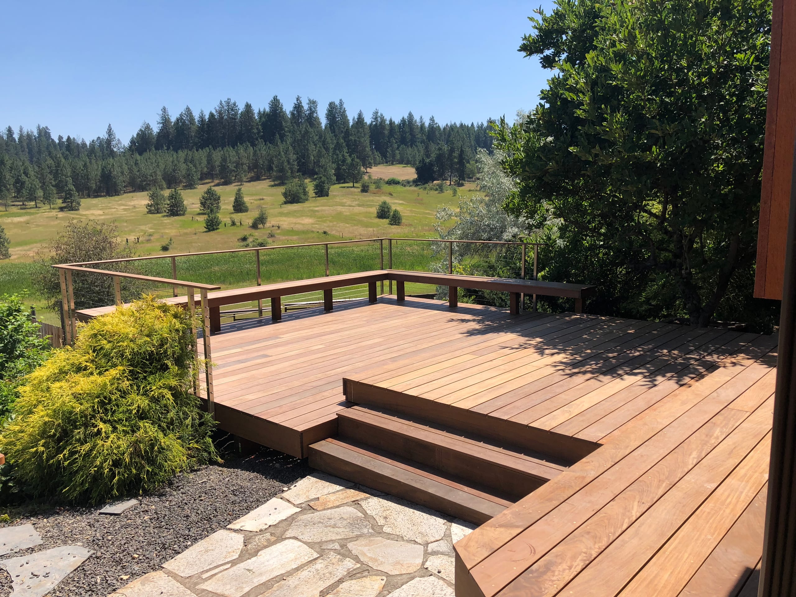 Reddish composite deck with wide steps and cable-style railing overlooking pasture, meadow and pines with flagstone path