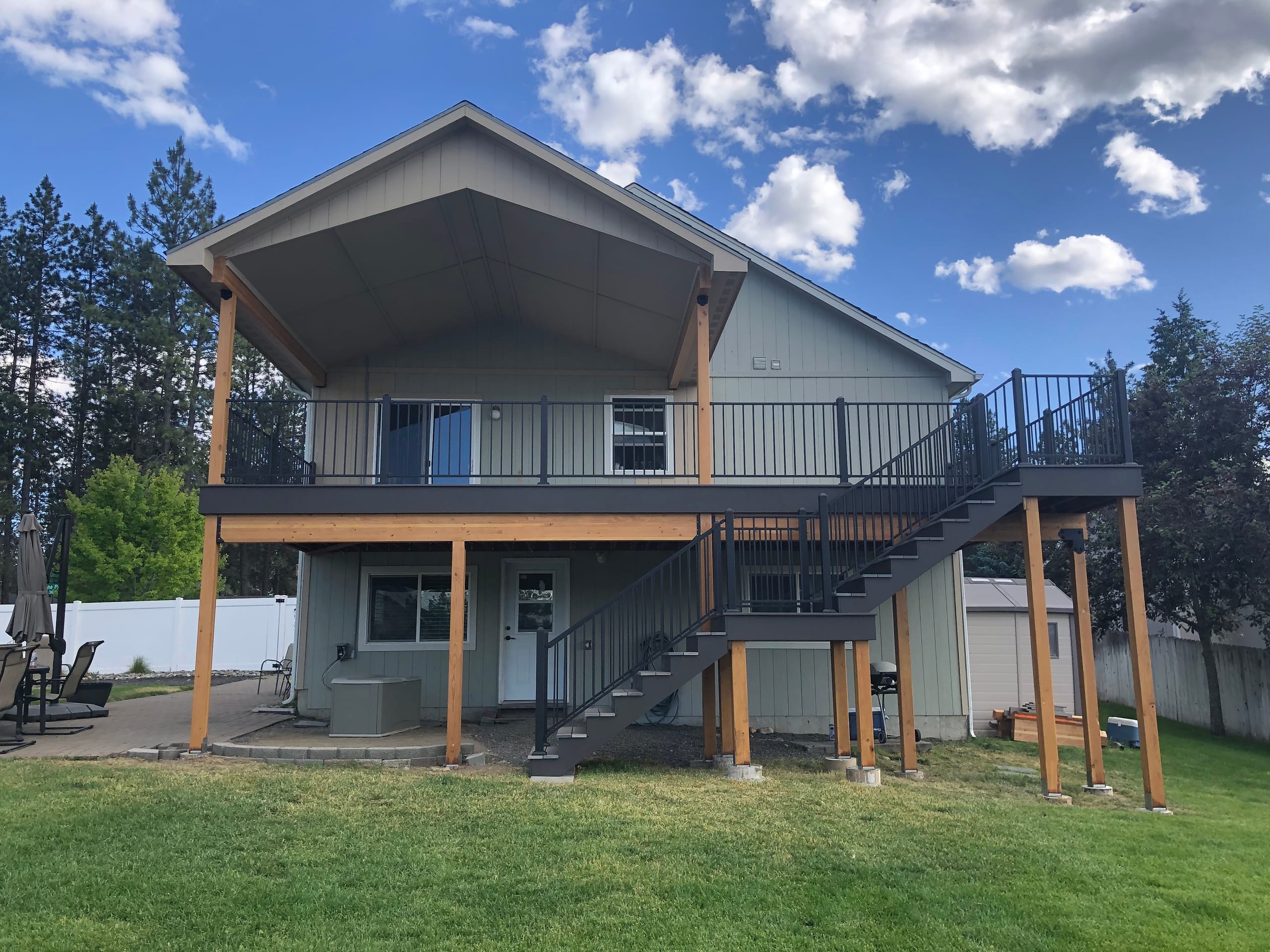 Sage house with elevated deck under gabled cover with cedar posts, black railing, stairs and lower patio, blue sky and lawn
