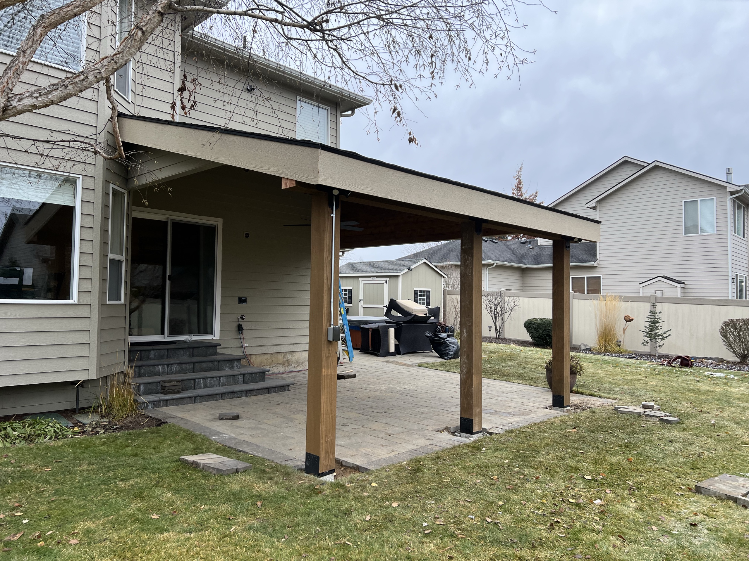 Shed-roof patio cover with timber posts over a paver patio behind a tan two-story house, dormant lawn
