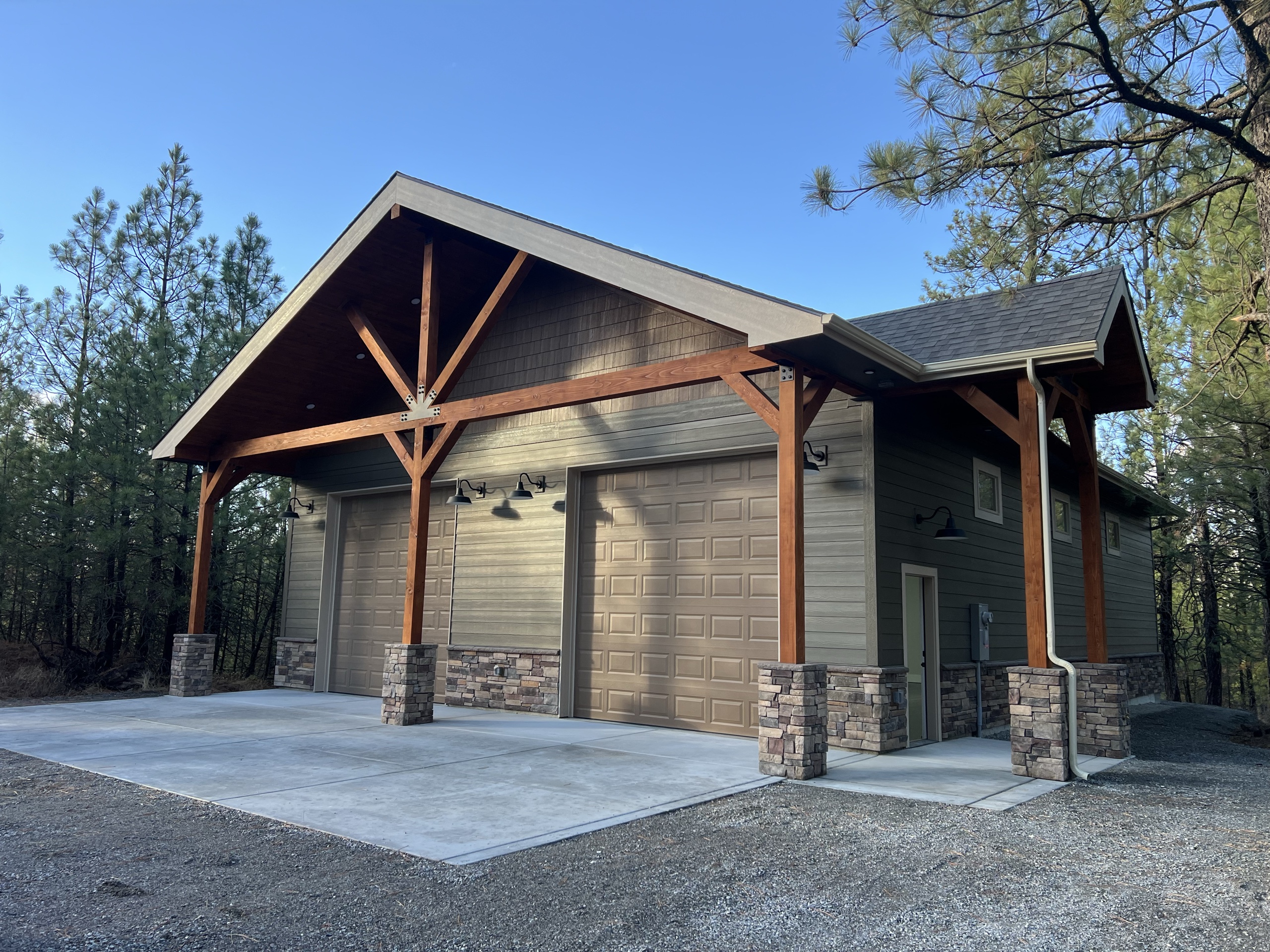 Shop building with timber-frame gable entry, stone pillars, two garage doors and concrete apron among pine trees, blue sky