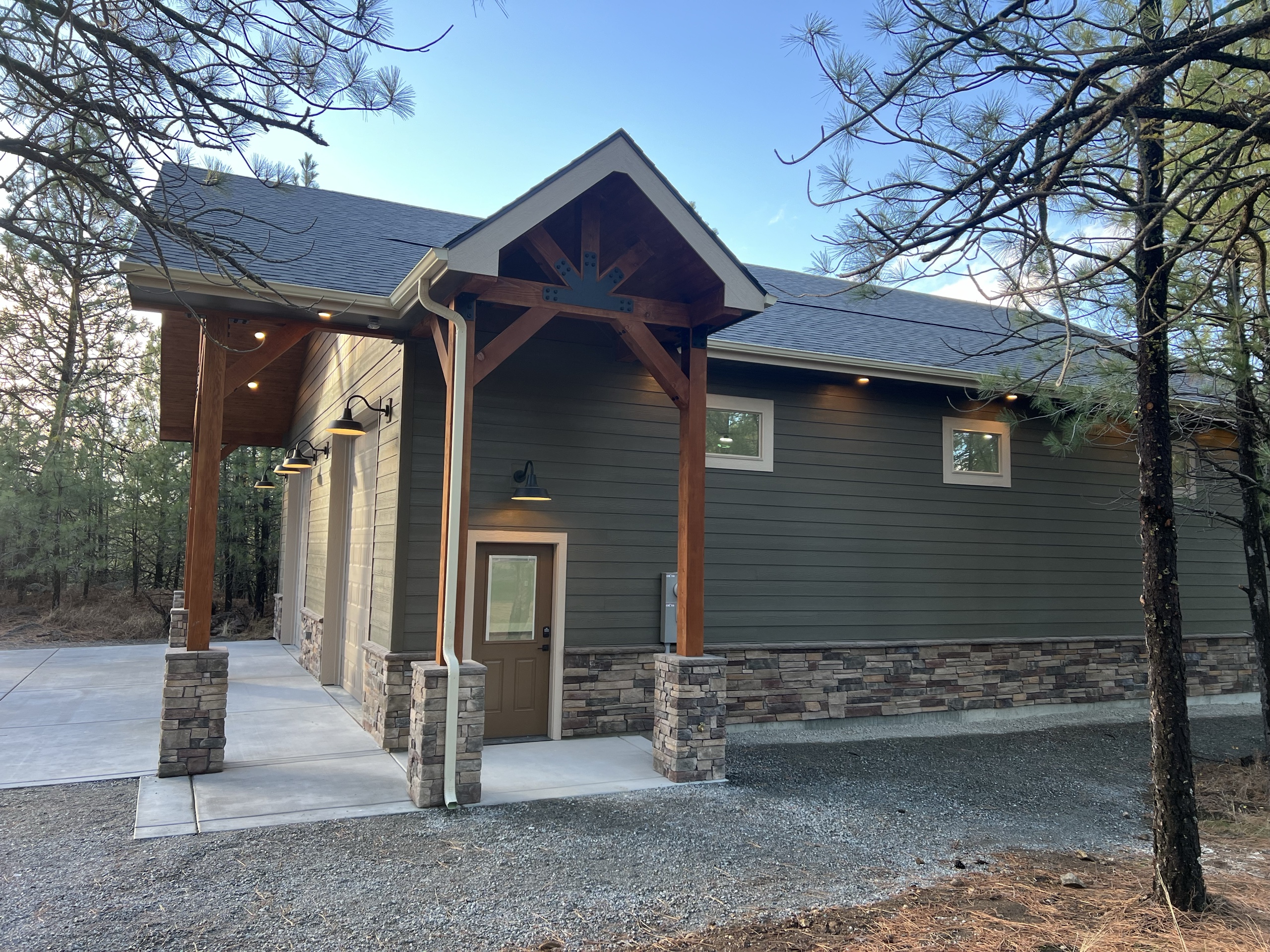 Shop building with a timber-frame gable entry on stone-wrapped posts and a stone wainscot among pines