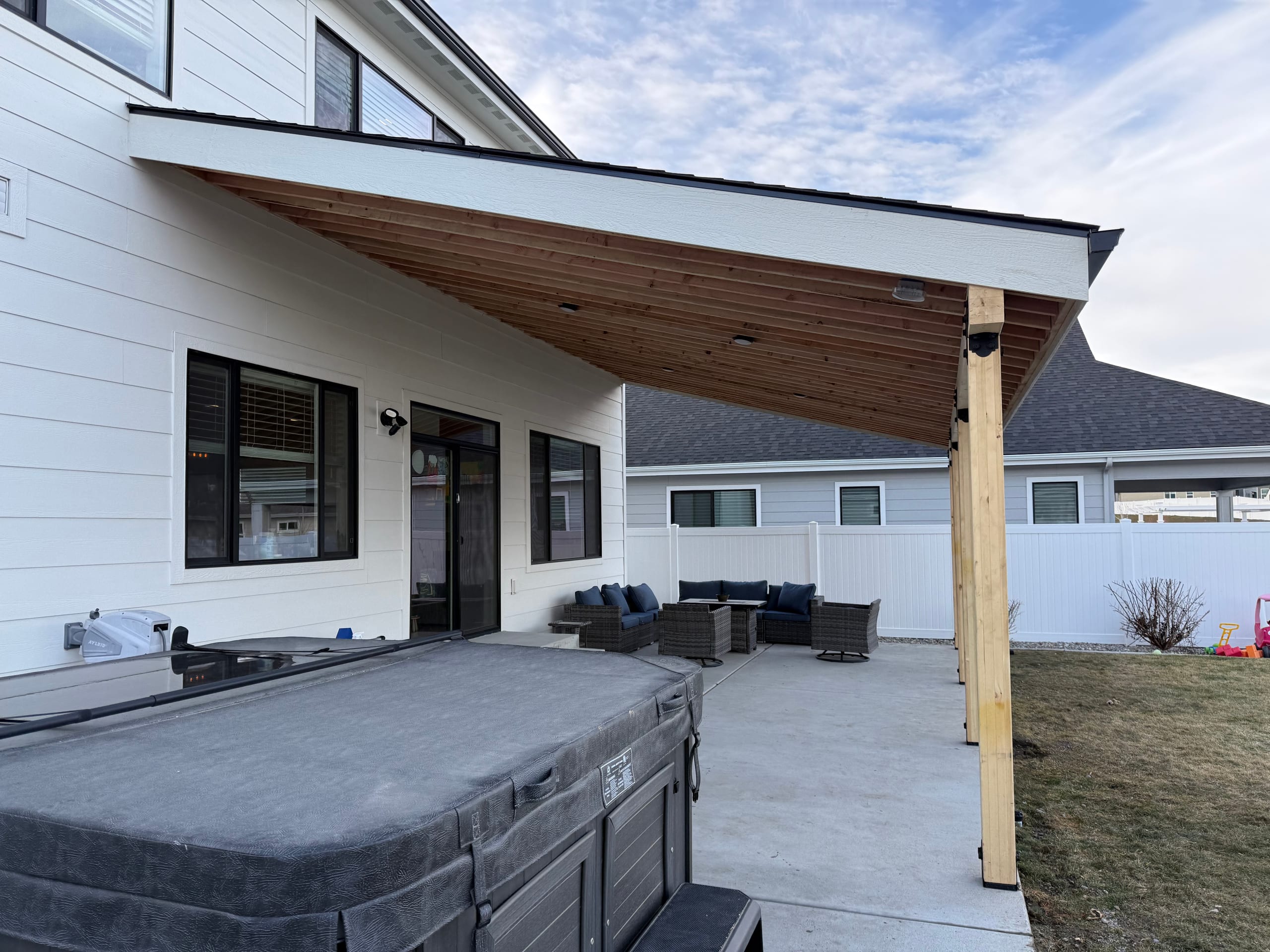Sloped stained-wood patio cover on a white two-story house over a concrete patio with hot tub and lounge furniture