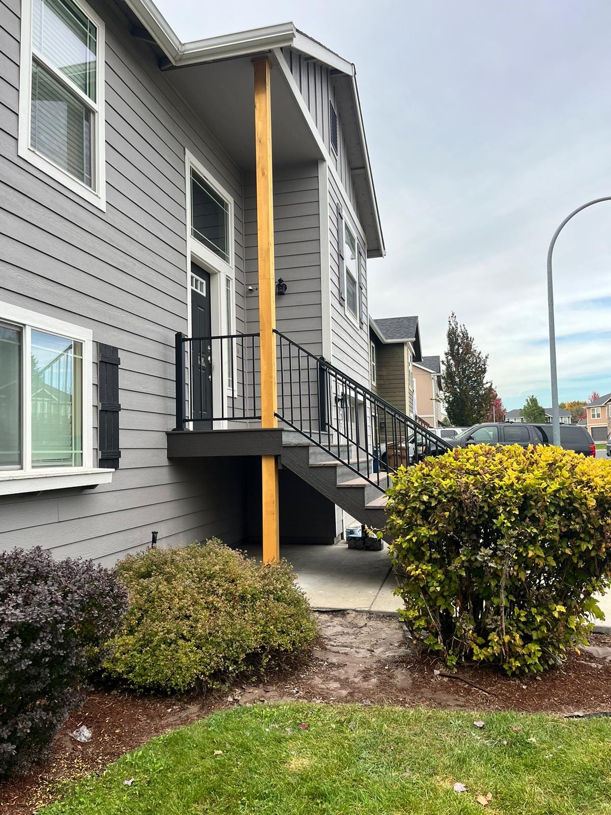 Small elevated entry landing with black railing and wood post on a grey two-story house, landscaped lawn