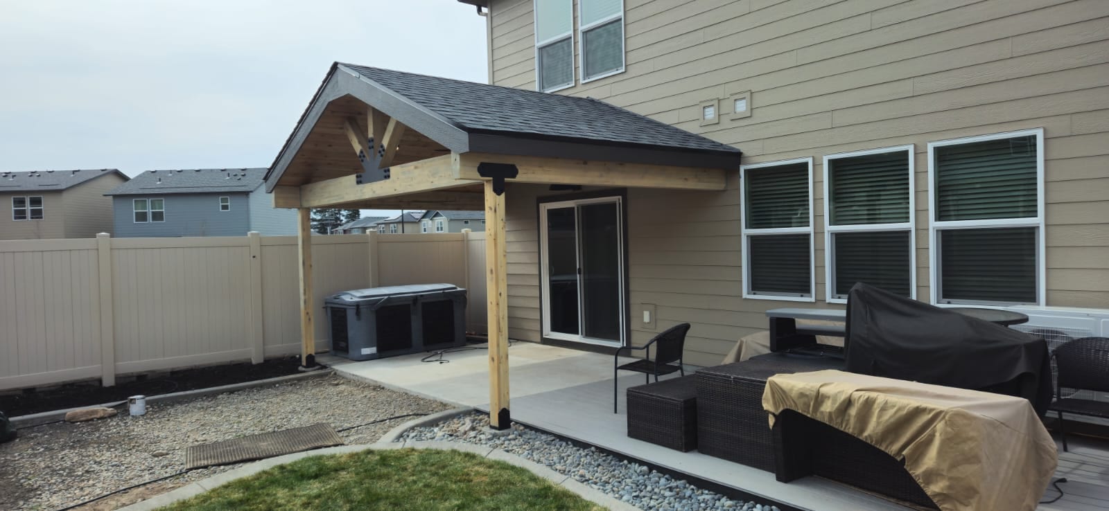 Small gable patio cover on a tan two-story house over a concrete patio with covered furniture and hot tub, vinyl fence