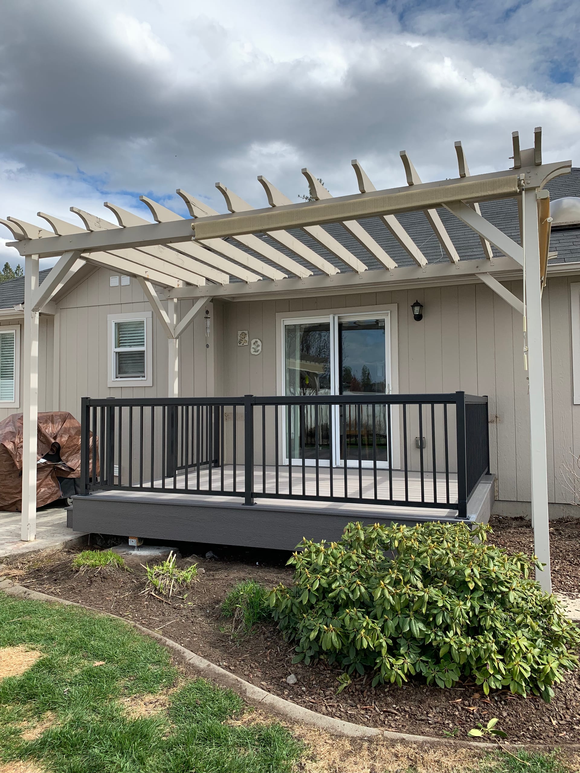 Small gray composite deck with black metal picket railing under a cream wood pergola at a tan house, landscaped beds