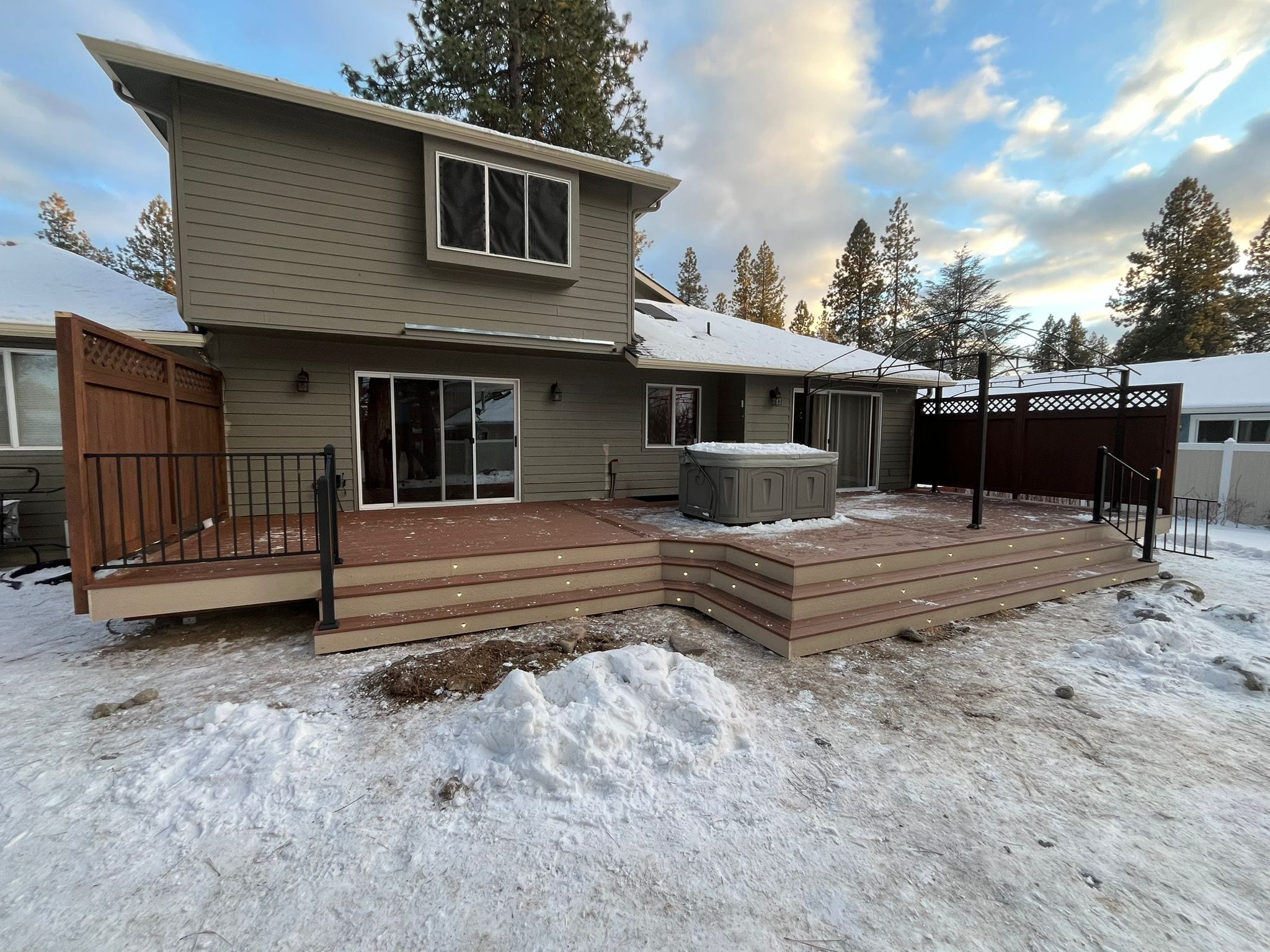 Snowy backyard with new multi-level gray composite deck with black railing, privacy panels and hot tub at tan house