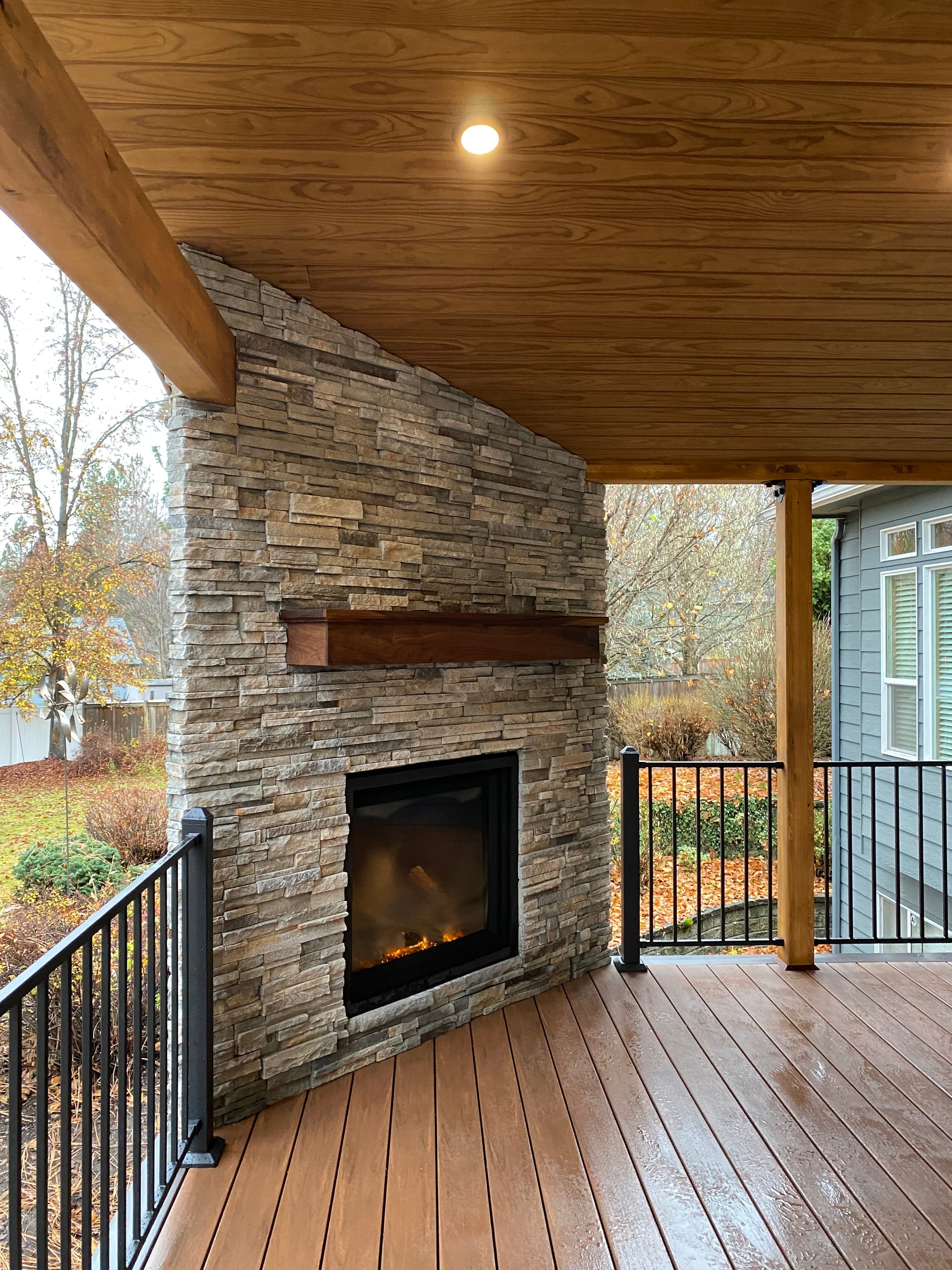Stacked-stone corner fireplace with wood mantel and glowing gas fire on a brown composite deck under a cedar ceiling