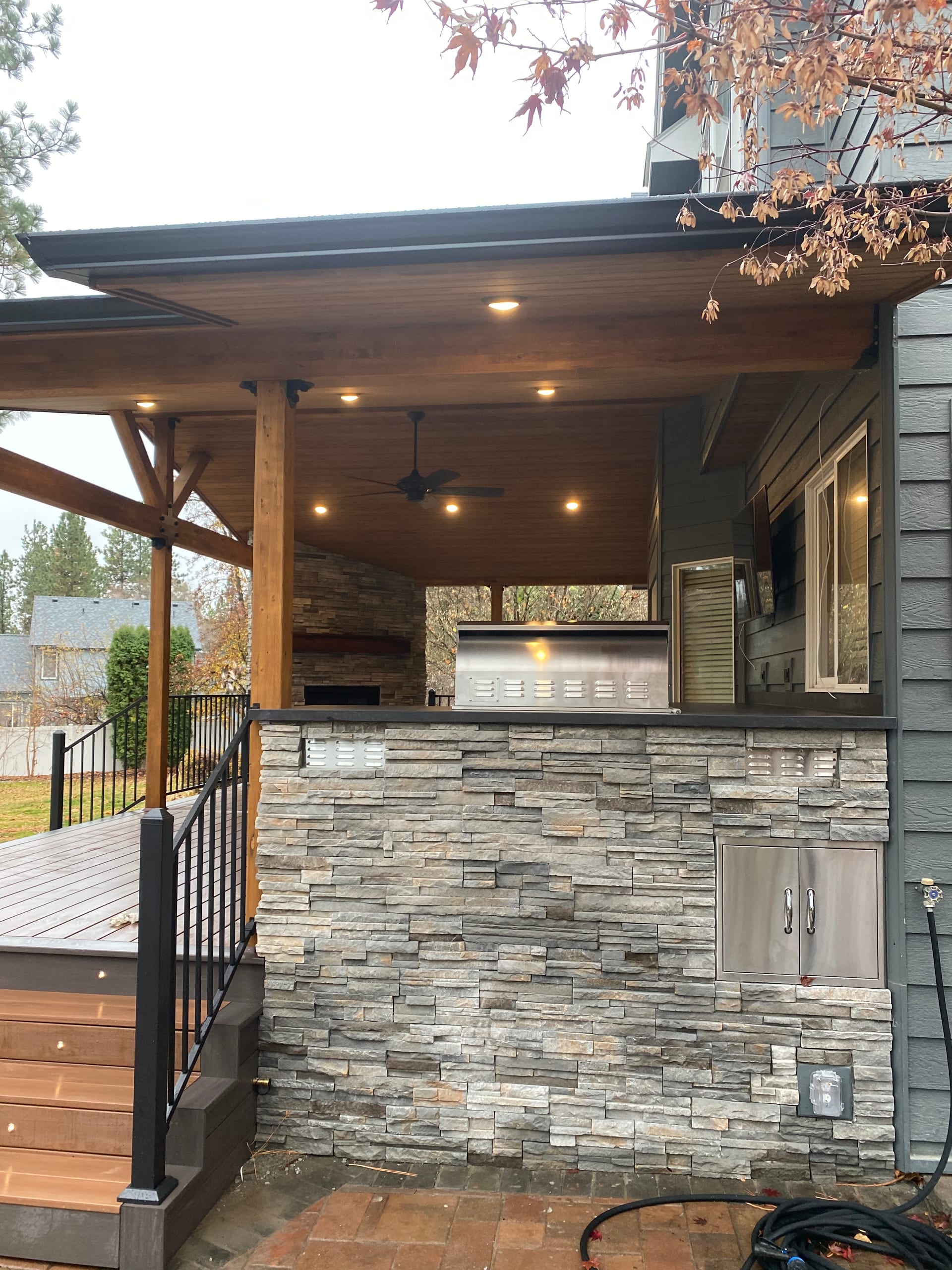 Stone-faced outdoor kitchen under a covered deck with timber post, recessed lighting, black railing, gray house