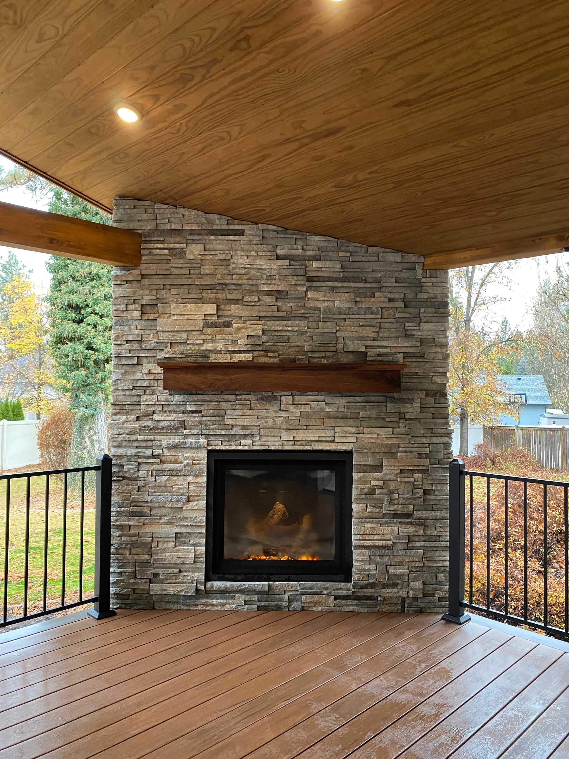 Stone fireplace with lit fire, wood mantel, cedar ceiling and black picket railing on warm composite deck