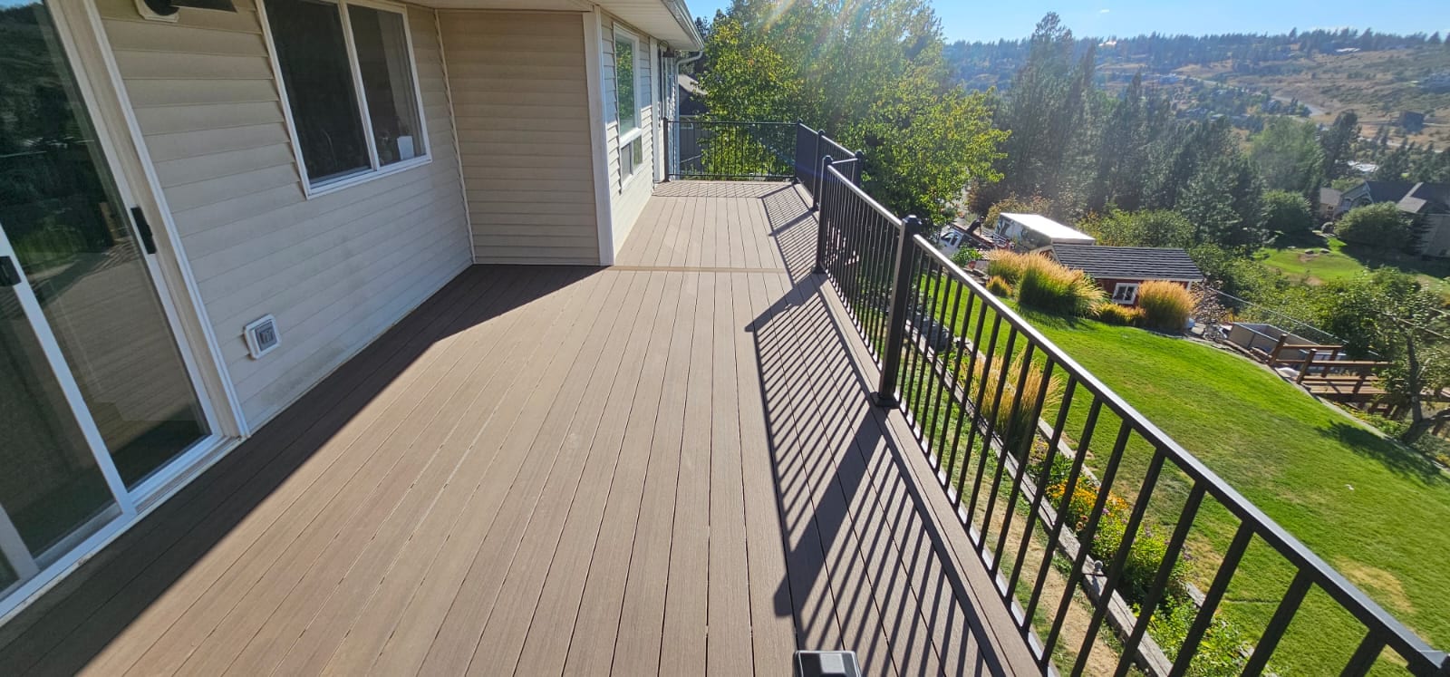Tan-brown composite deck along house with sliding doors, black metal picket railing and sunny hillside view