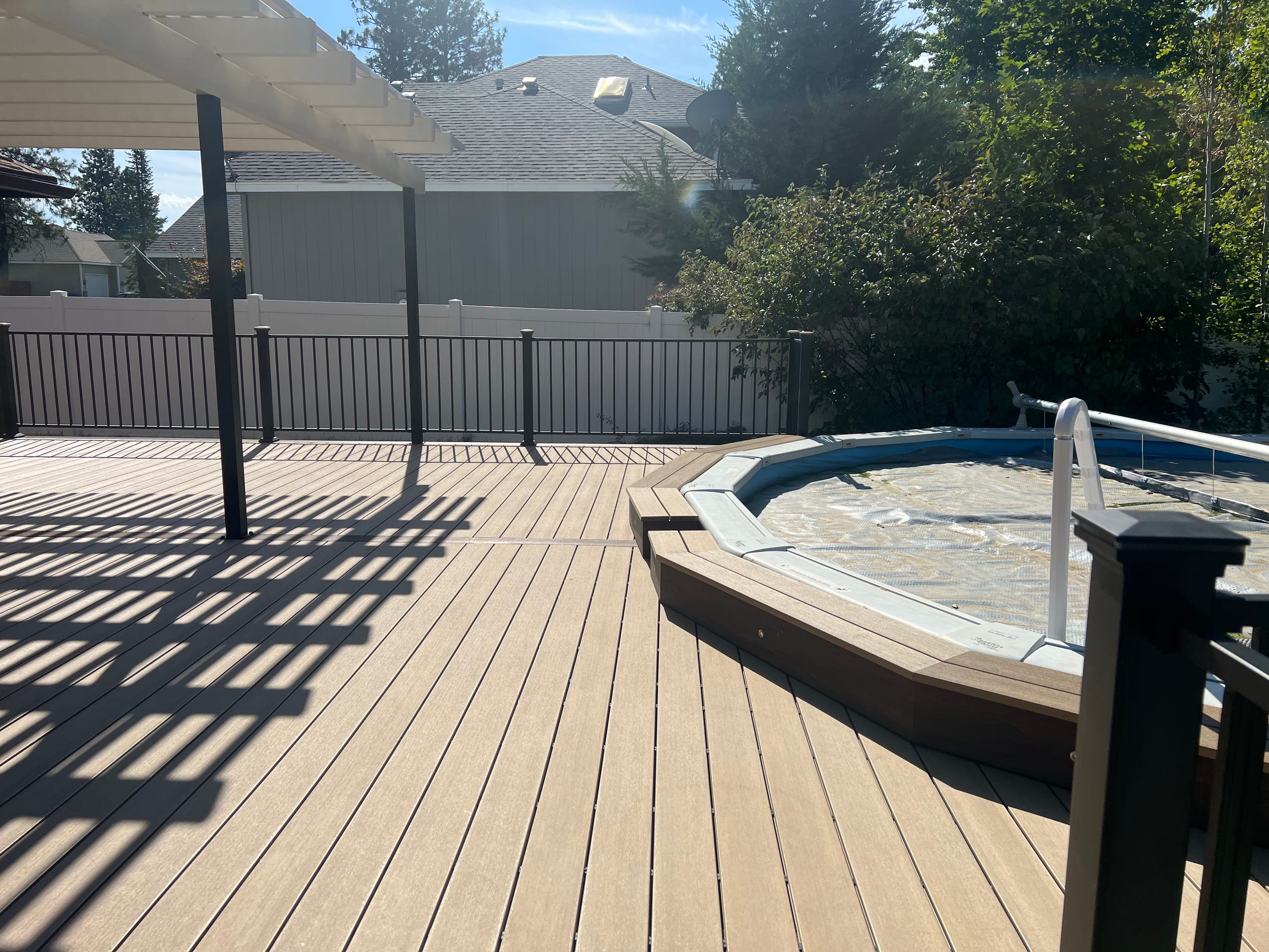 Tan composite deck beneath pergola with octagonal pool surround and grey vinyl-style fence