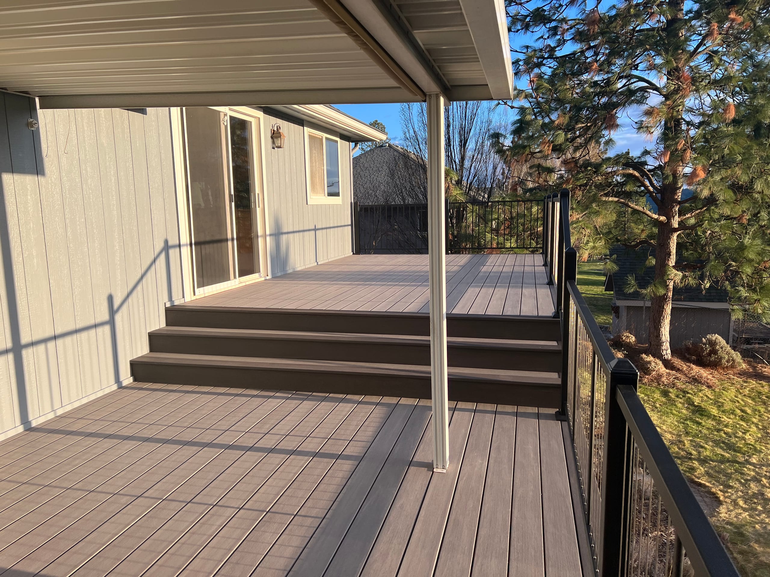 Tan composite deck with brown stair steps under aluminium patio cover and black glass-panel railing