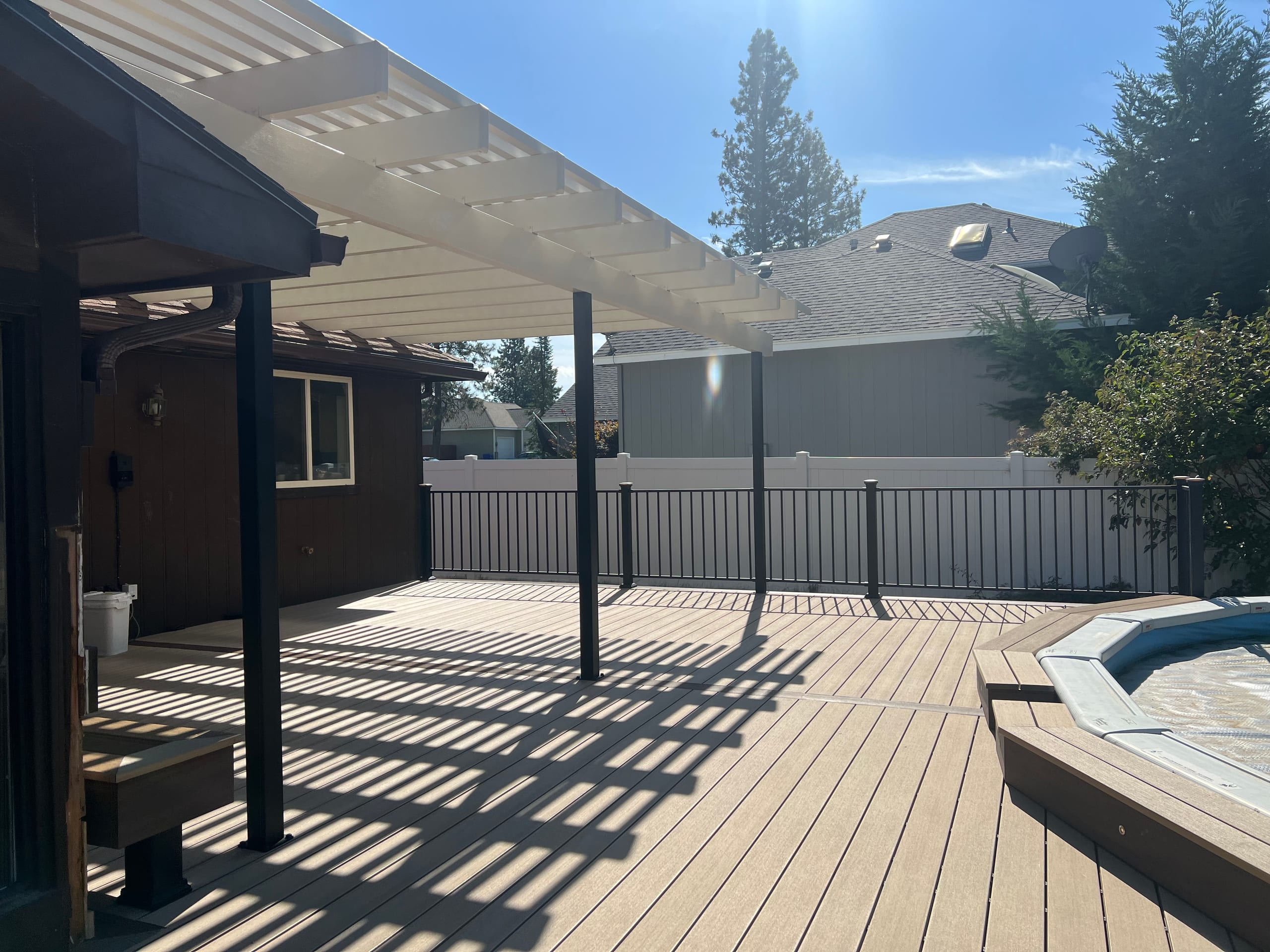 Tan composite deck under a white louvered pergola next to a brown house, with pool surround at right