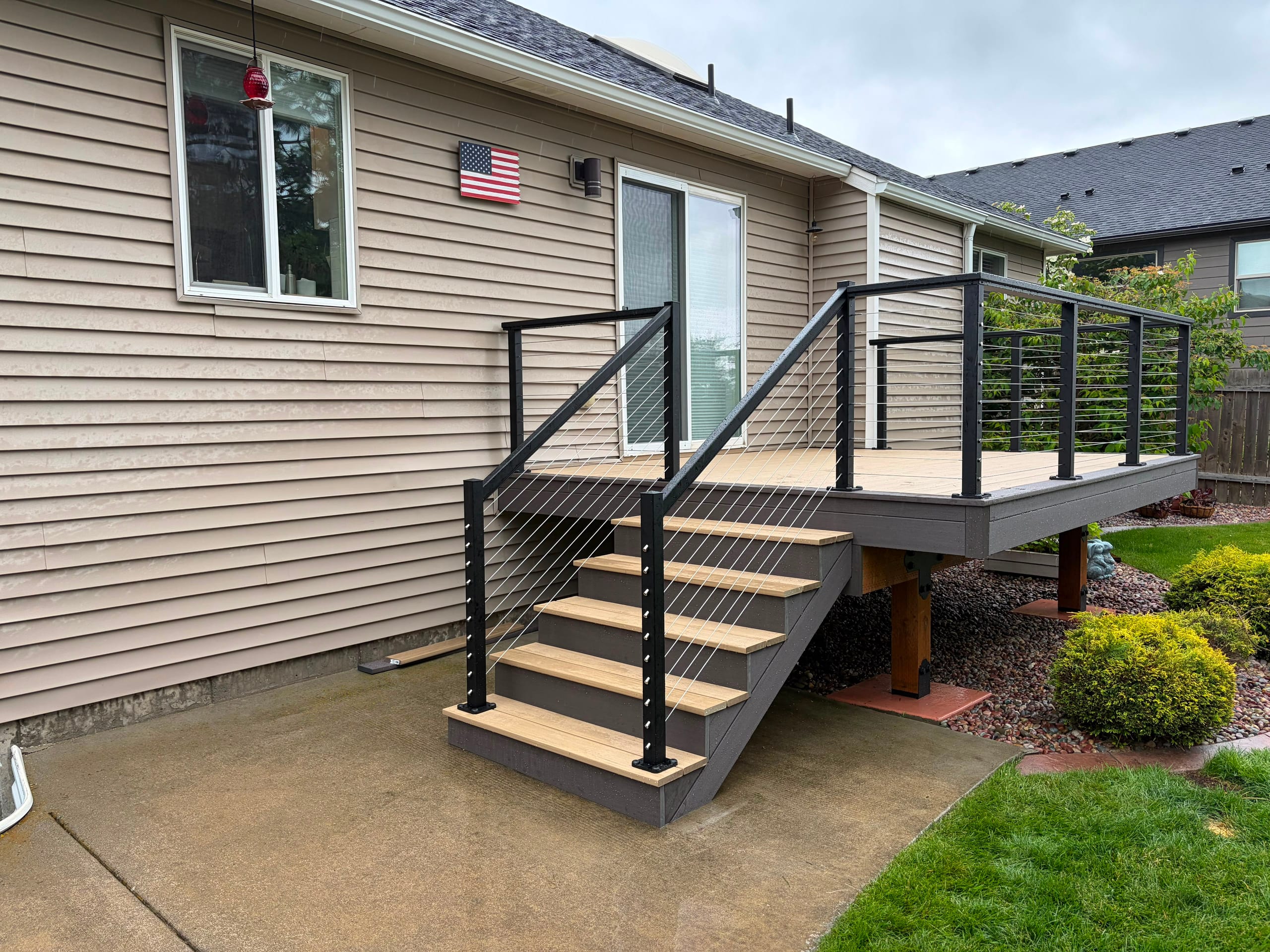 Tan composite stairs with black cable railing and grey fascia leading down to concrete patio beside tan house