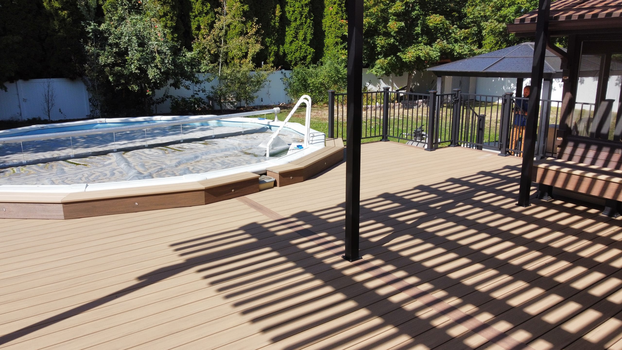 Tan deck surrounding an above-ground pool, with pergola shadows across the boards