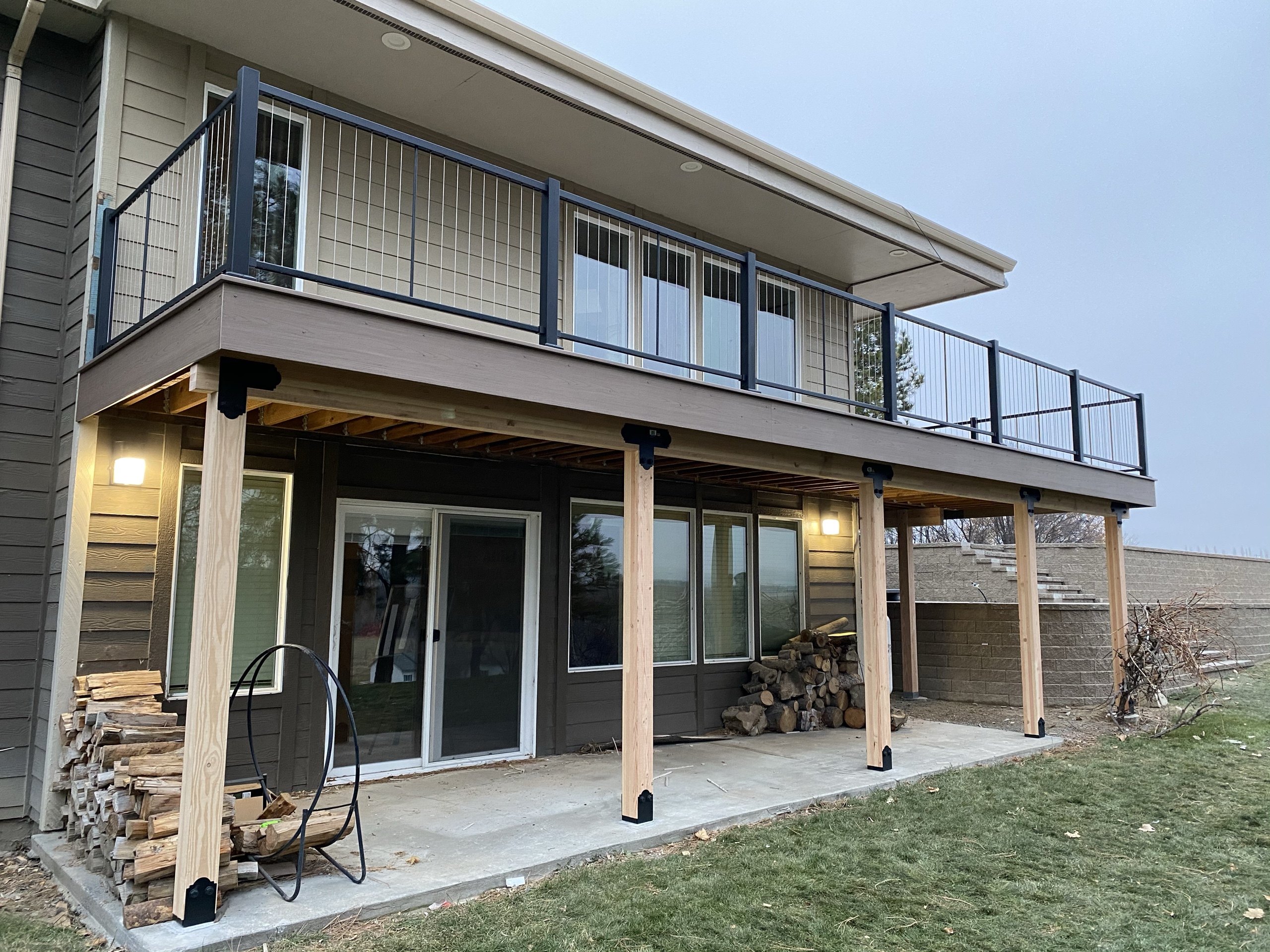 Tan house with elevated cable-railing deck on cedar posts, covered lower concrete patio with firewood, evening lights