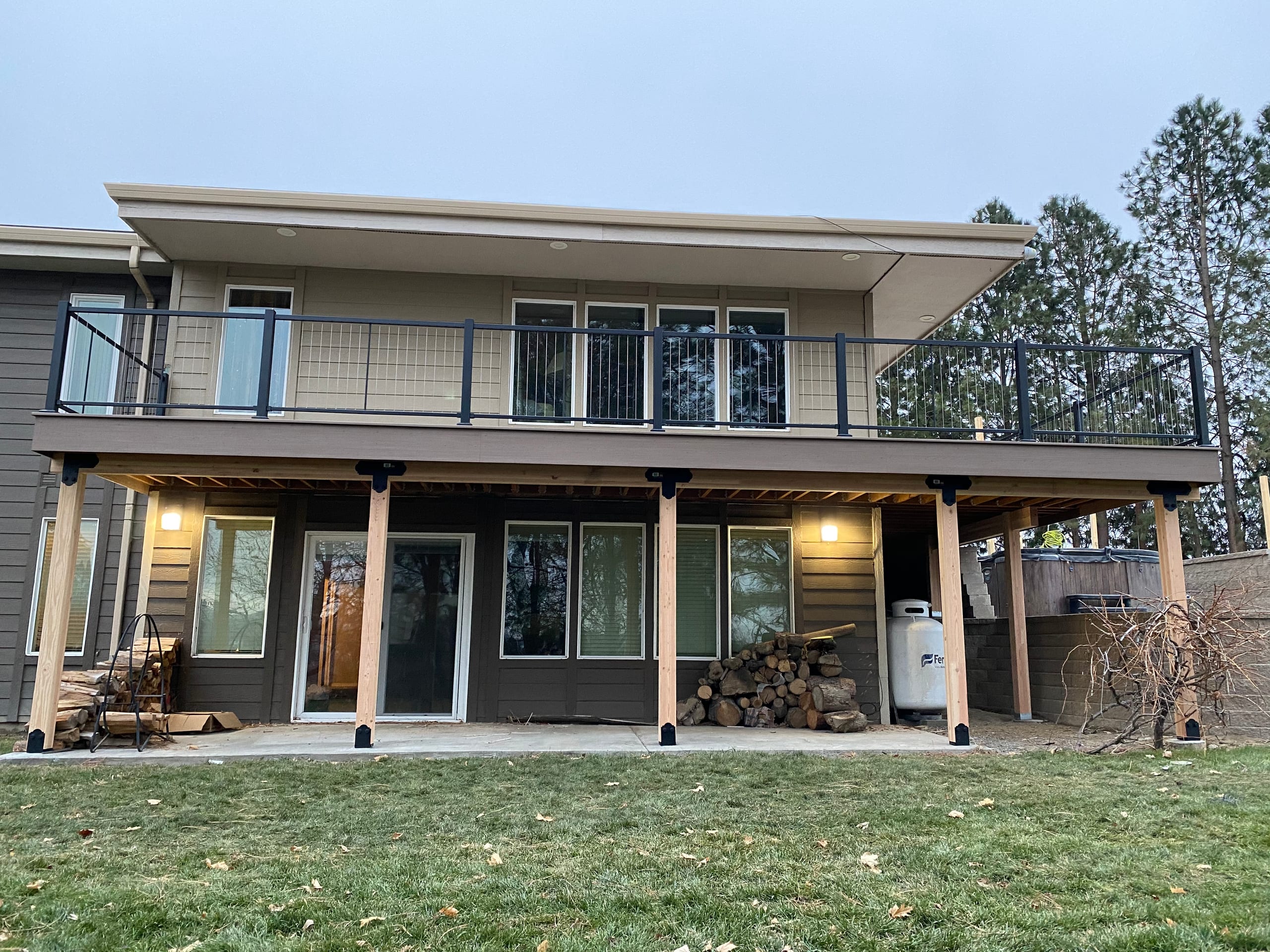 Tan house with elevated deck with cable railing and cedar posts, lit lower patio, firewood and propane tank, green lawn
