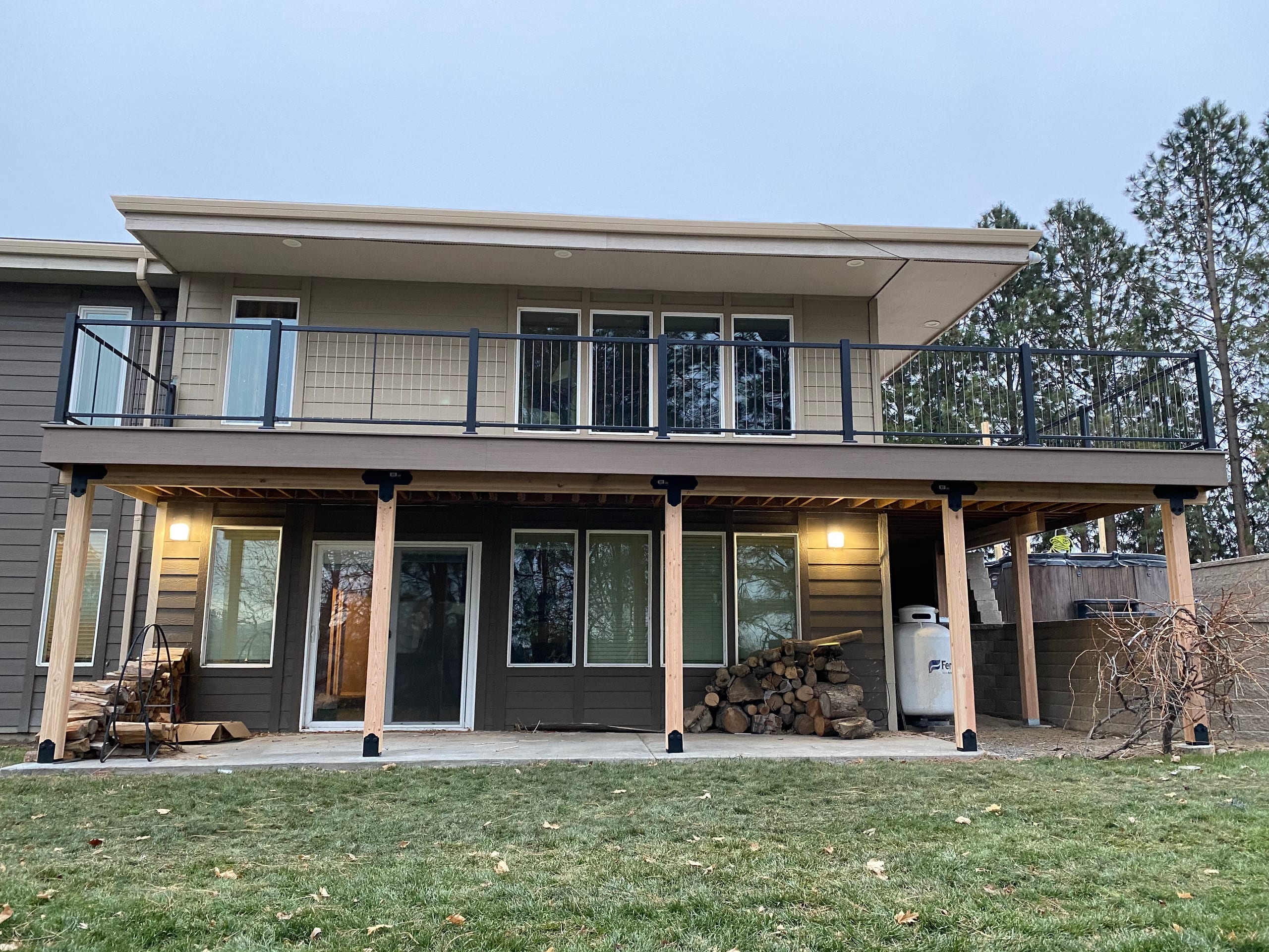 Tan house with elevated deck with cable railing on cedar posts, lit lower patio and lawn