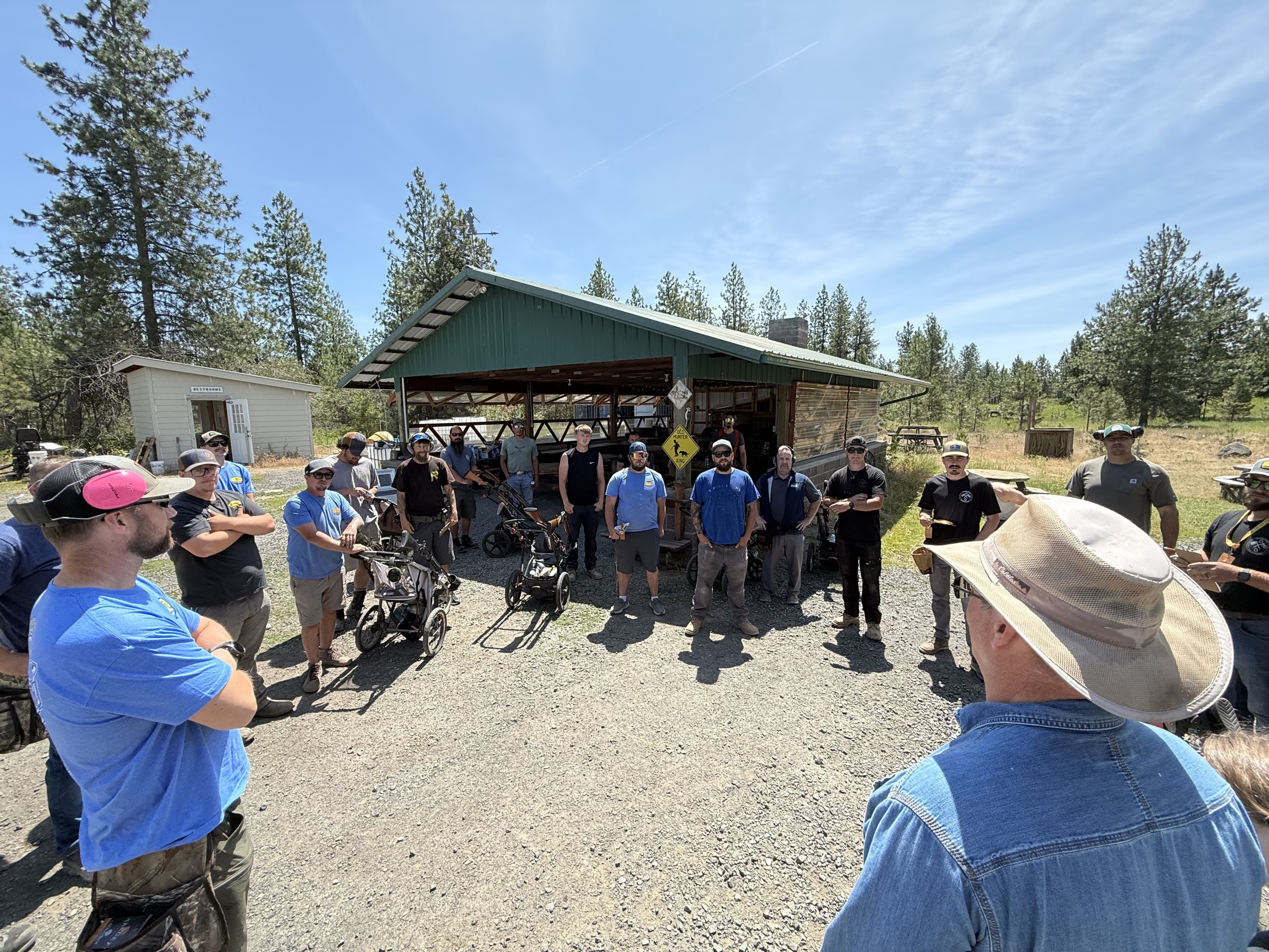 Team gathering outdoors at a gravel yard in front of a green-roofed open shed, men in blue shirts and hats listening, sunny