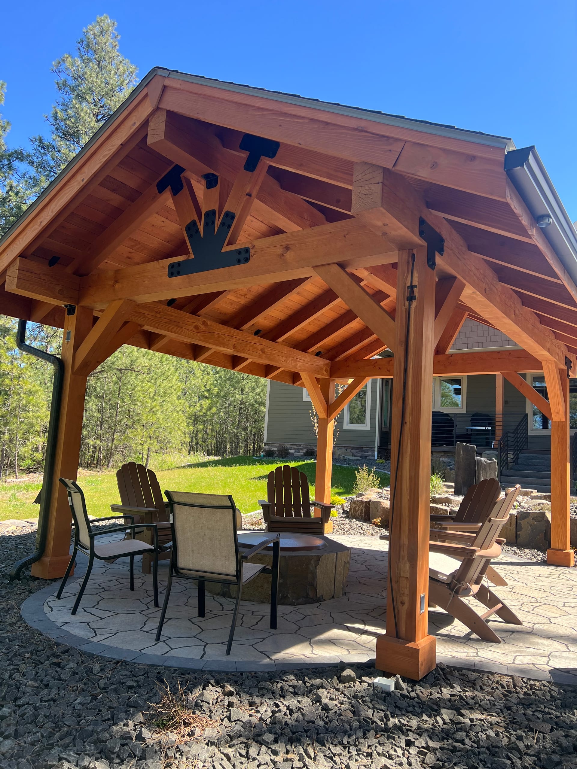 Timber-frame cedar-look pavilion with gable truss details over paver patio with firepit and chairs, blue sky, pines