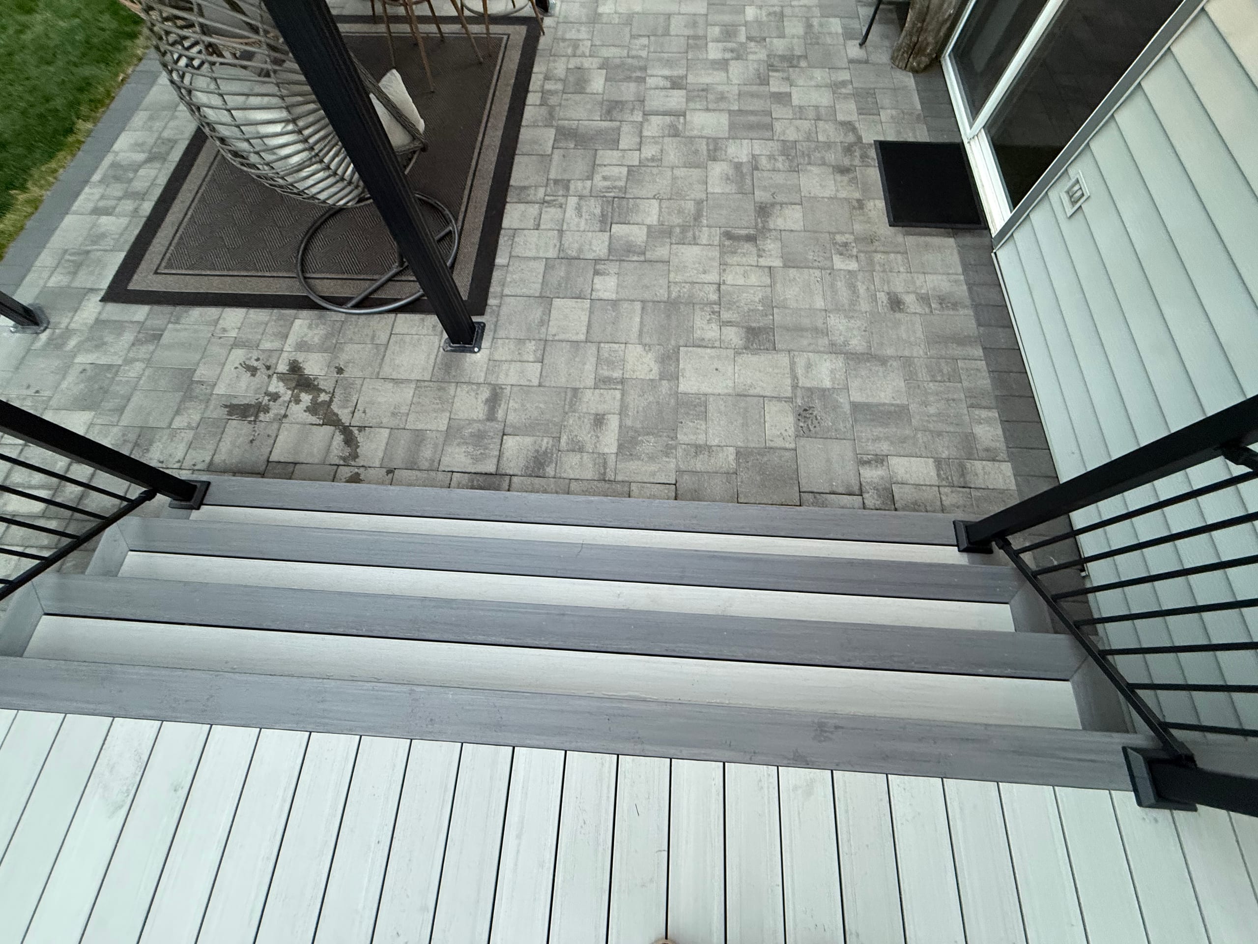 Top-down of light grey composite deck with stairs to a grey paver patio, black railing and outdoor rug