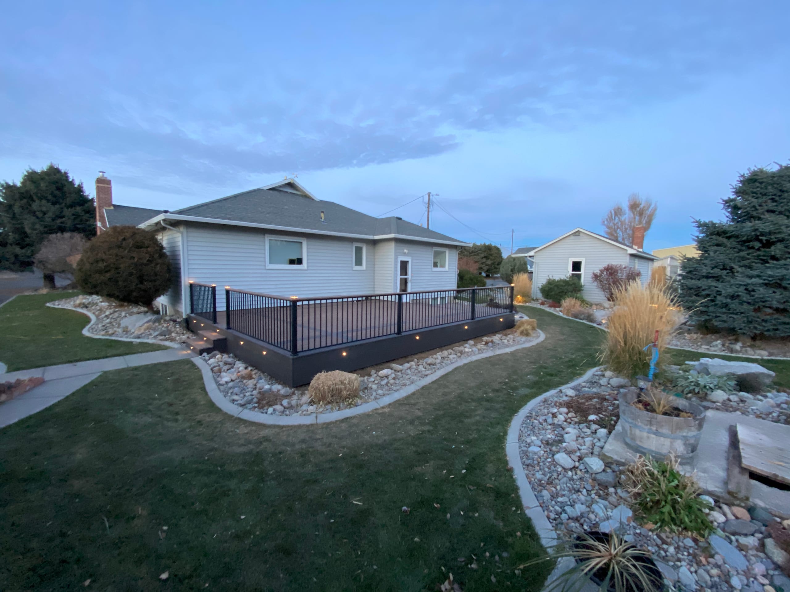 Twilight backyard view of low composite deck with black railing and fascia lights, curved rock edging, lawn