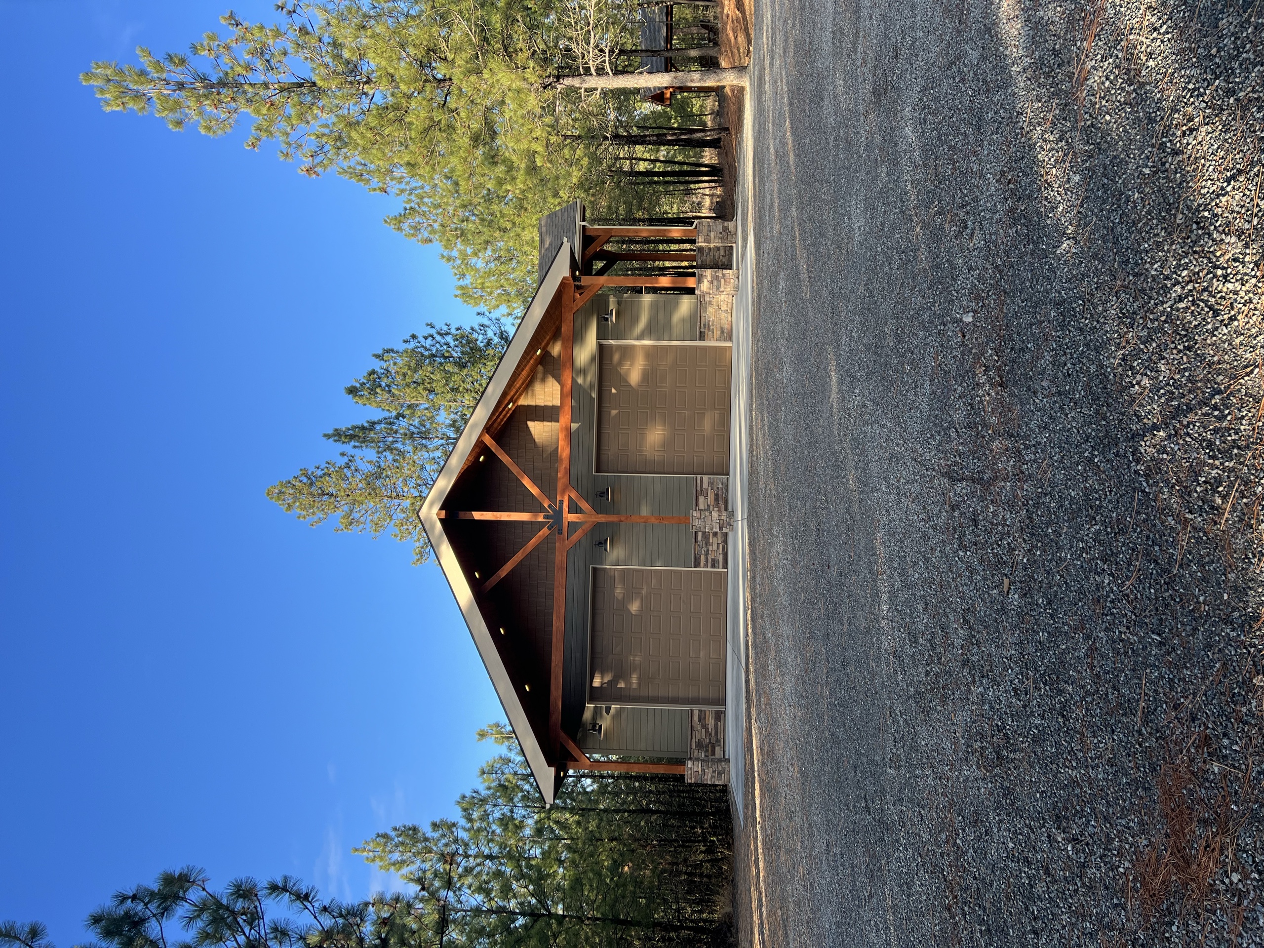 Two-door shop with a timber-frame gable entry set among tall pines on a gravel apron