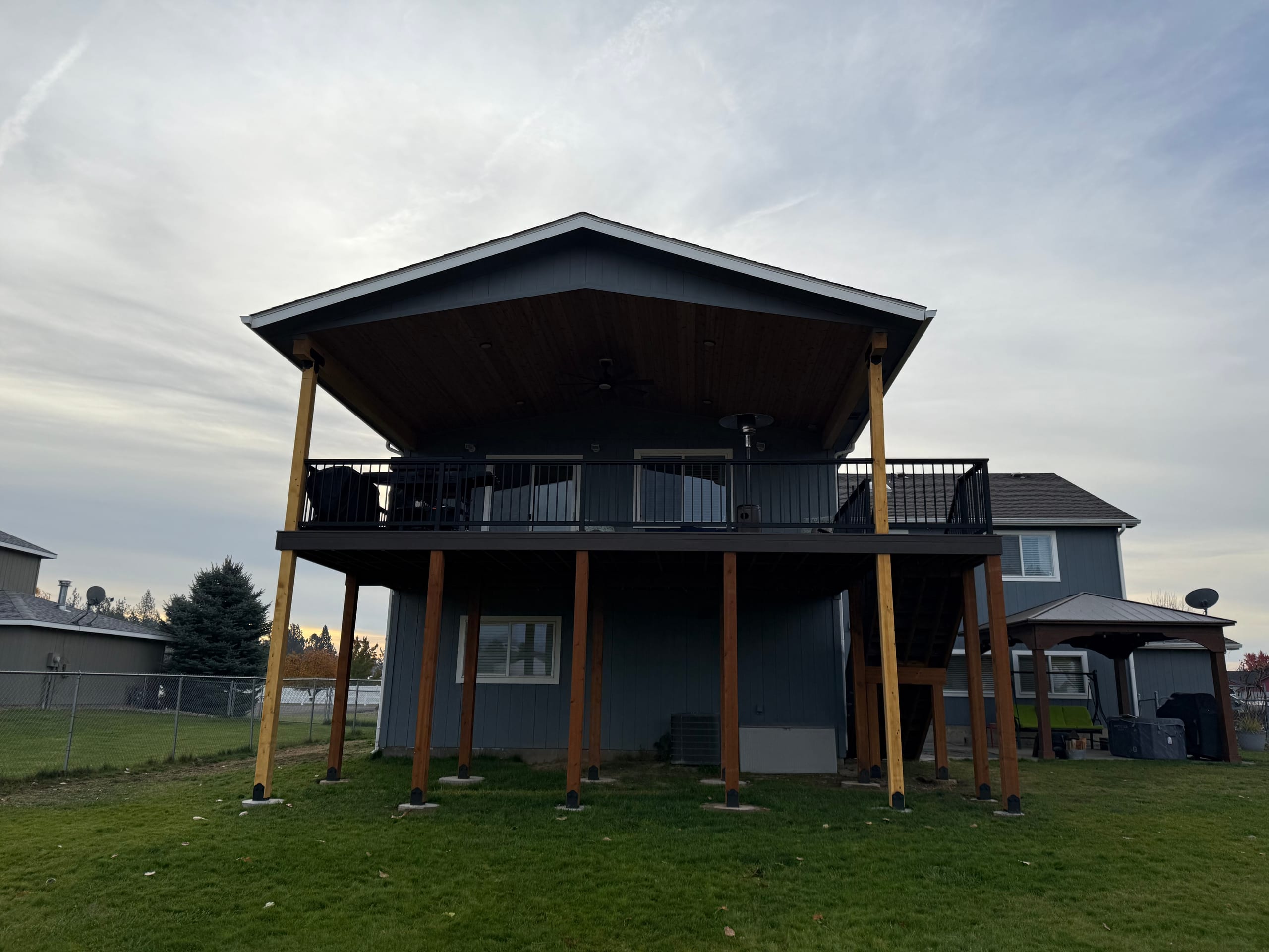 Two-level covered deck on dark gray house with timber posts, black railing, overcast sky, grass lawn