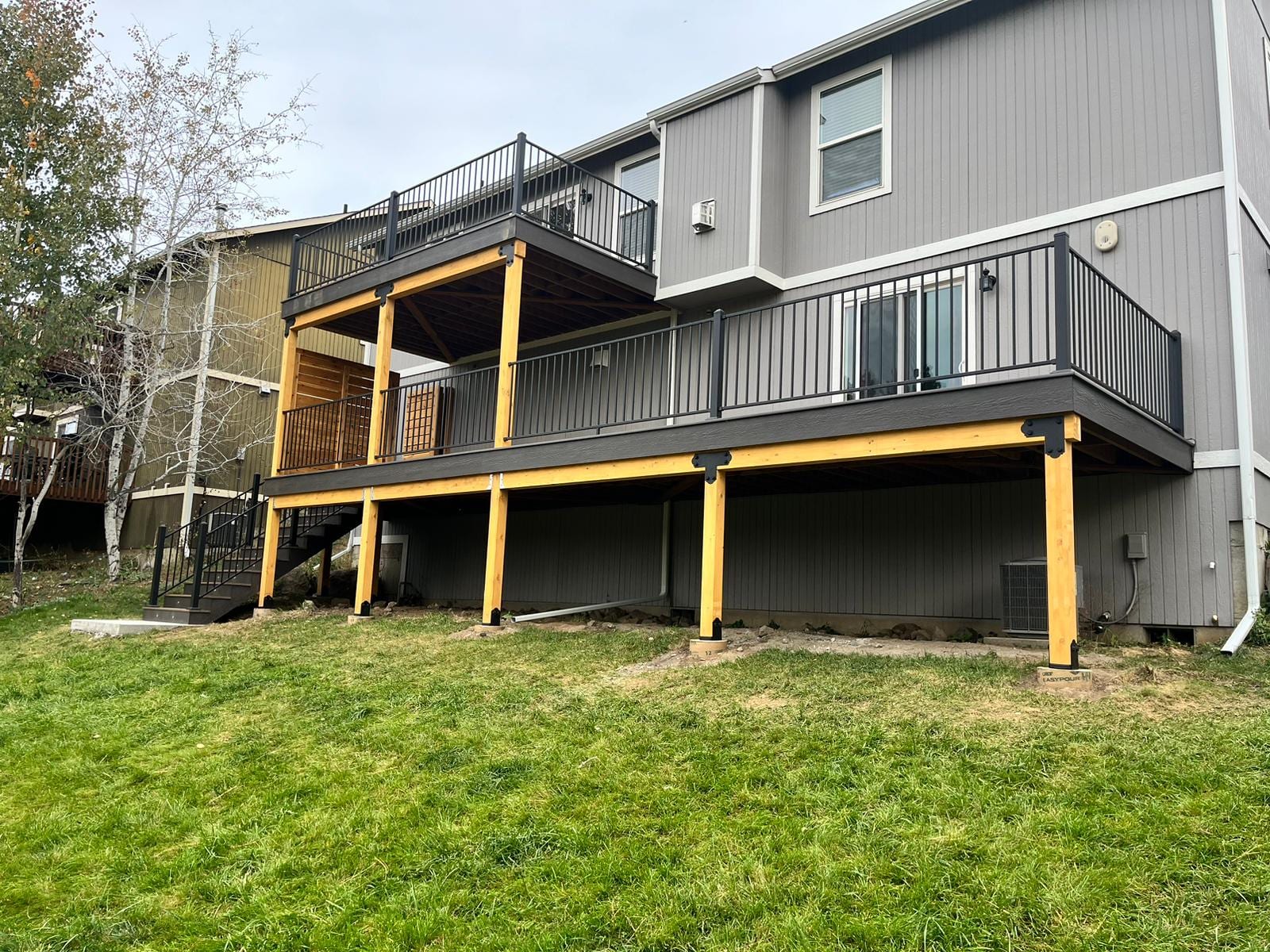 Two-level dark gray composite deck on yellow-stained posts with black railing and cedar privacy screen at a gray house