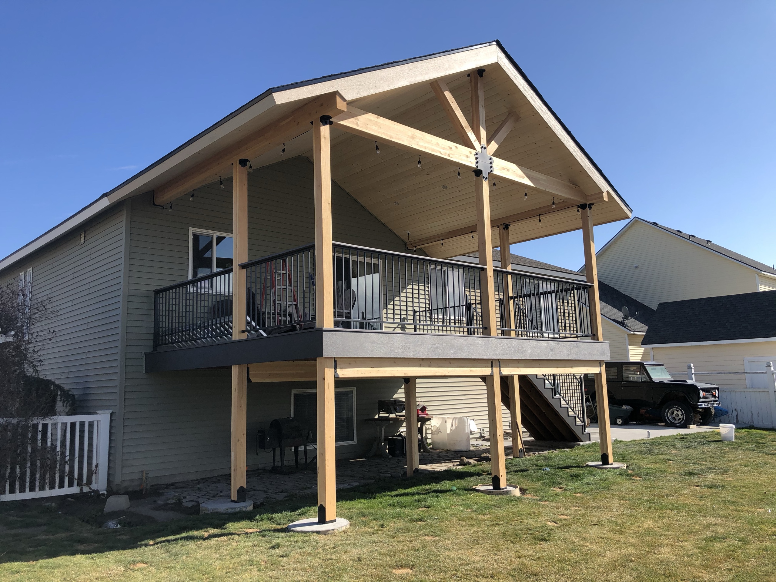 Two-level elevated deck with gable roof cover and king-post truss, black picket railing, yellow-stained posts, blue sky