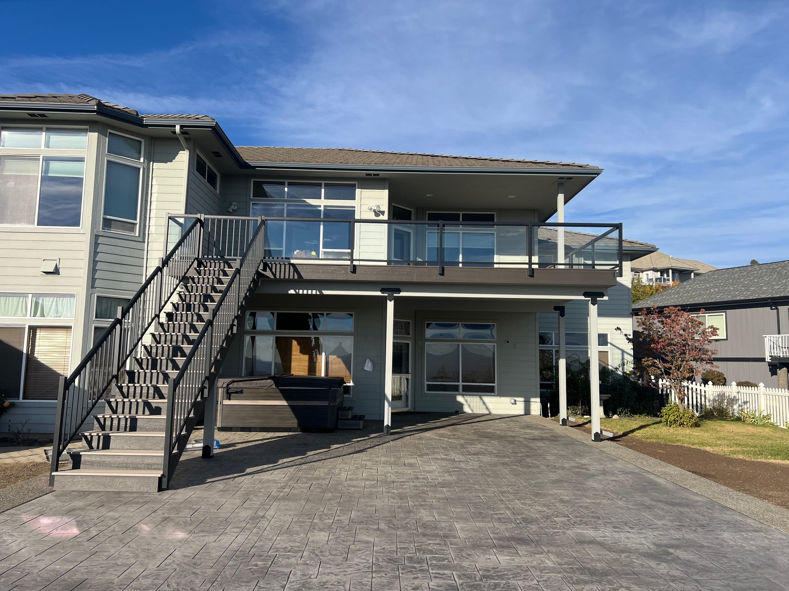 Two-story home with elevated deck with glass railing and black metal stairs over stamped concrete patio and hot tub underneath
