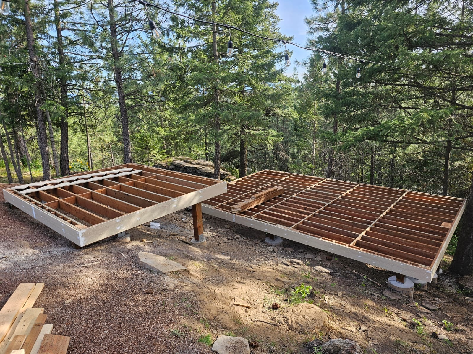 Two wood joist frames on concrete footings in a forest clearing before decking, lumber stacked nearby
