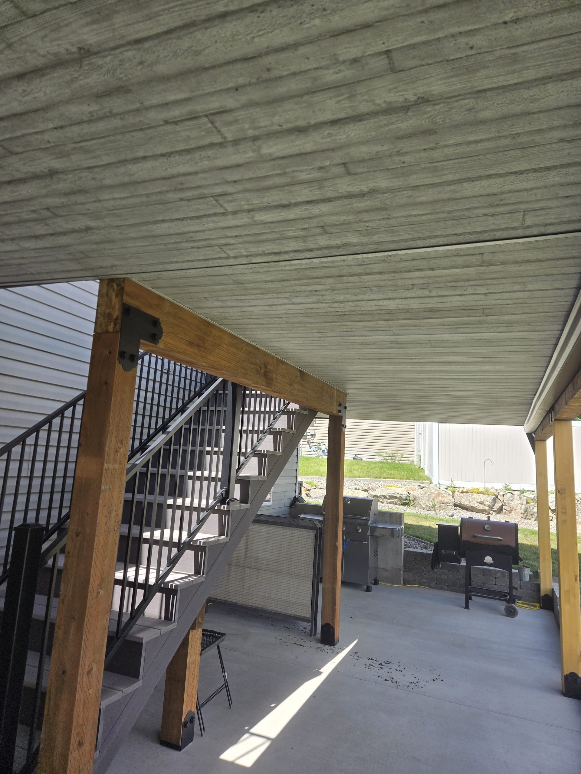 Underdeck ceiling with cedar posts and metal stair railing over concrete patio with outdoor kitchen and grill beyond