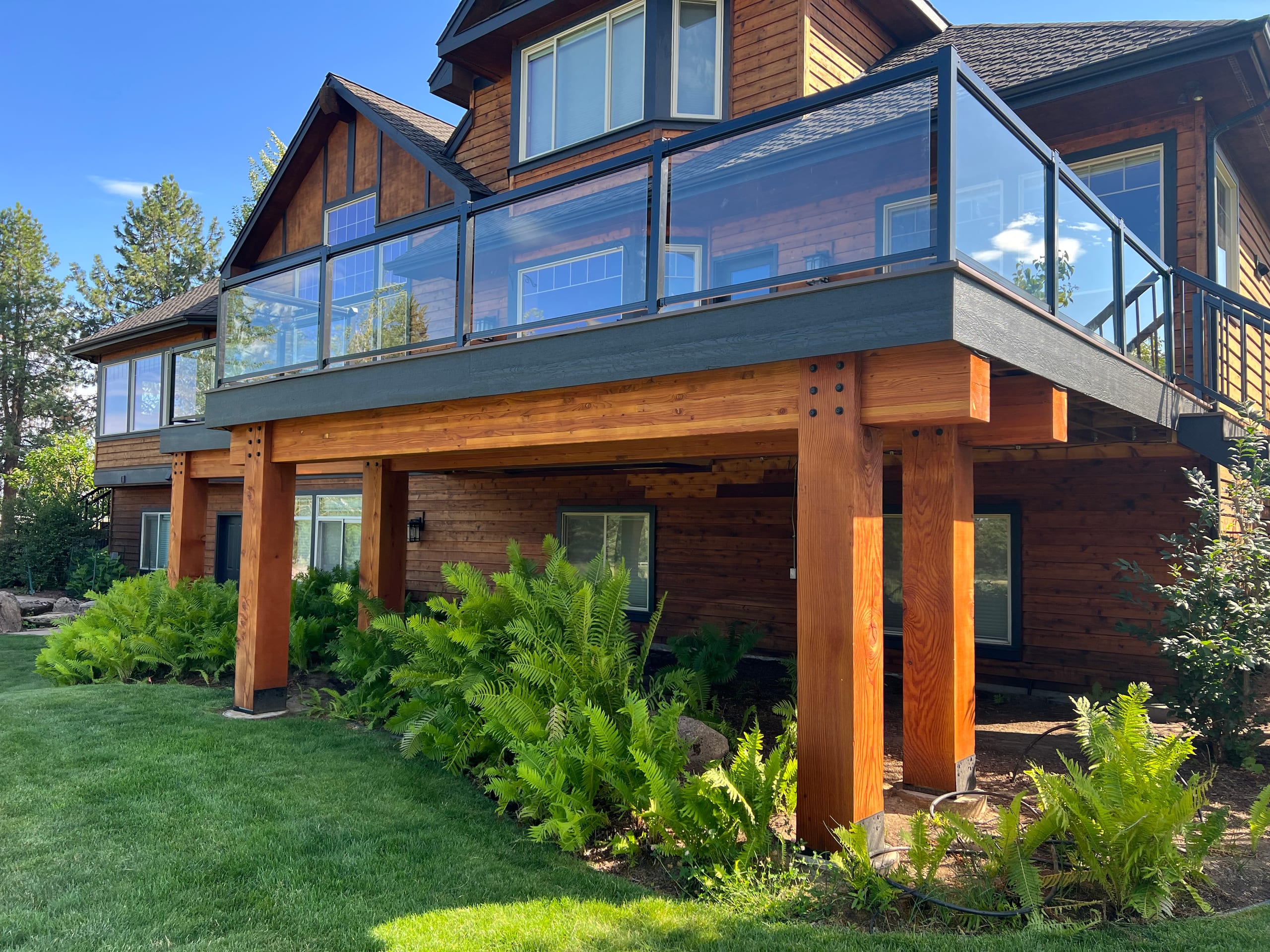 Underside of elevated deck with glass railing supported by large timber posts above ferns and lawn