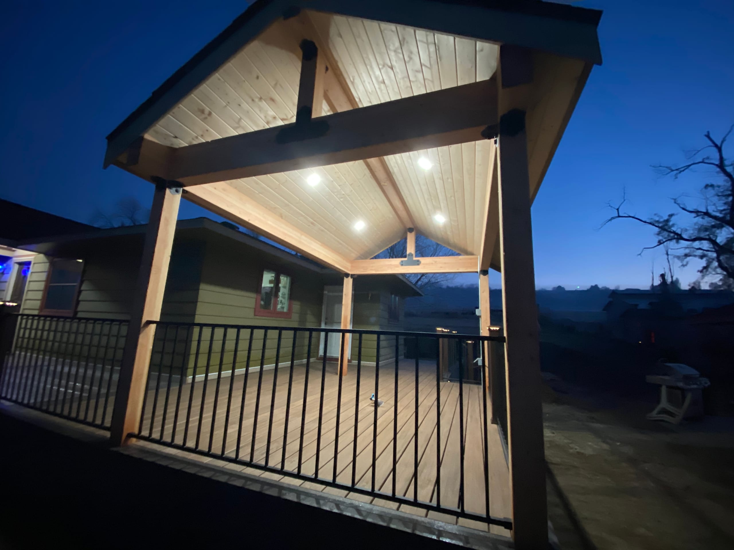 Underside of gable roof over deck with lit cedar ceiling and recessed lights, black railing, deep blue dusk sky