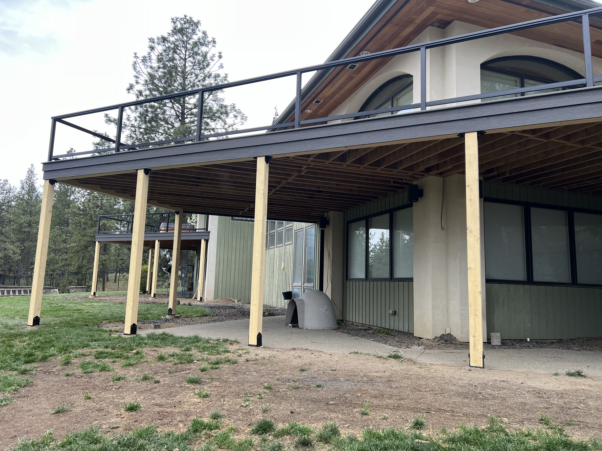 Underside of large elevated deck with exposed joists and tall timber posts, black railing above, dirt patio below