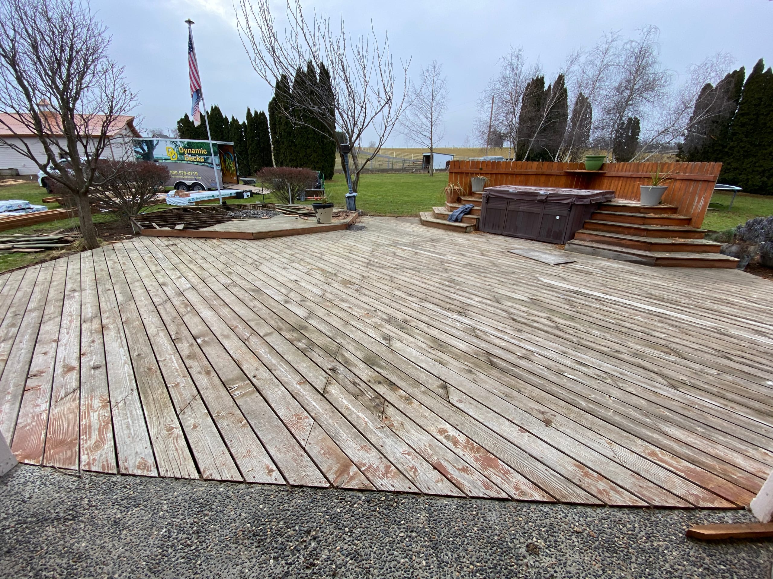Very large weathered grey wood deck with raised hot-tub platform in the distance, gravel edge, flag and rural field