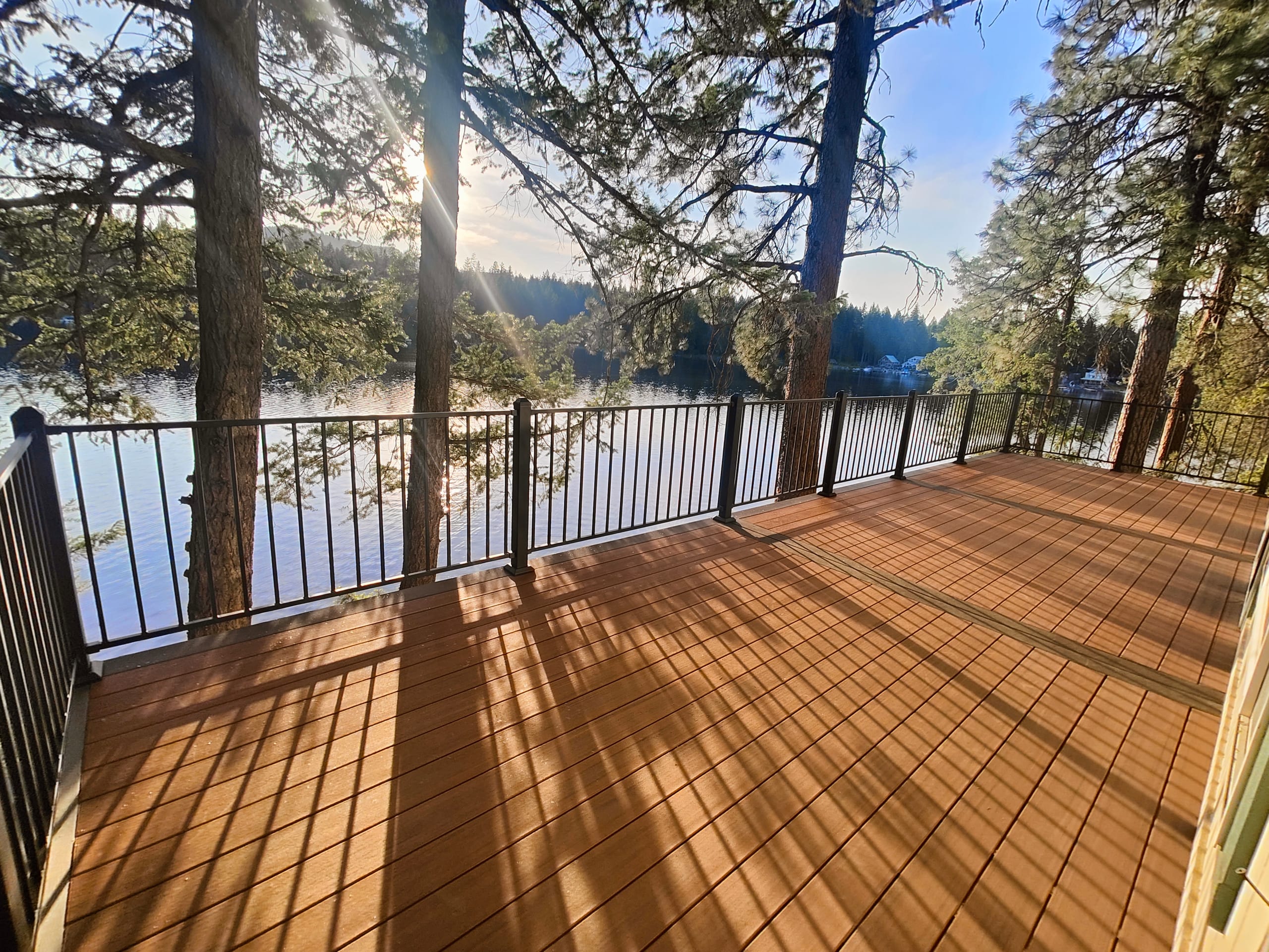 Warm cinnamon composite deck with black metal picket railing and long sun shadows overlooking a lake framed by pines