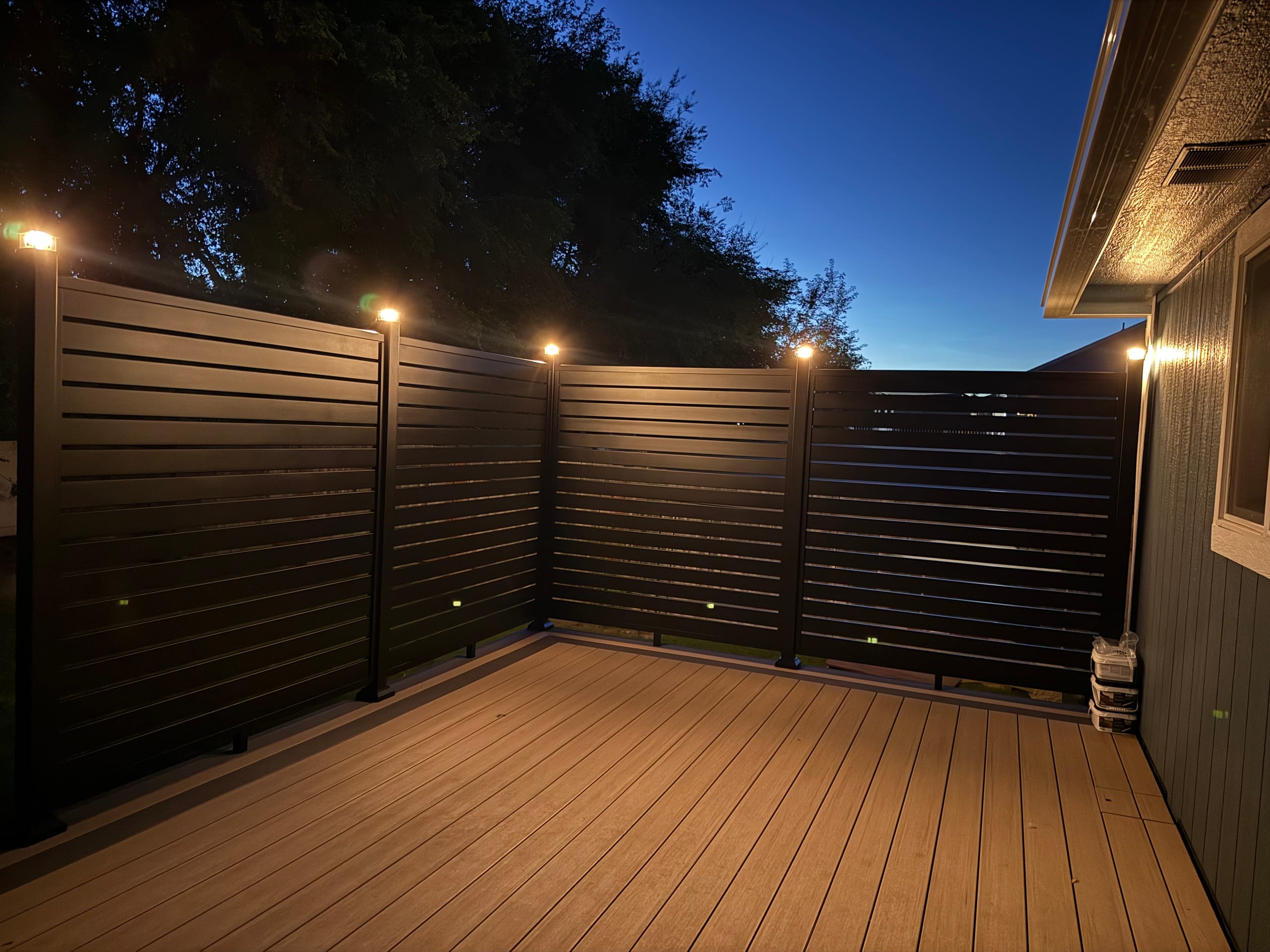 Warm-toned composite deck at dusk enclosed by dark horizontal-slat privacy wall with post-cap lights