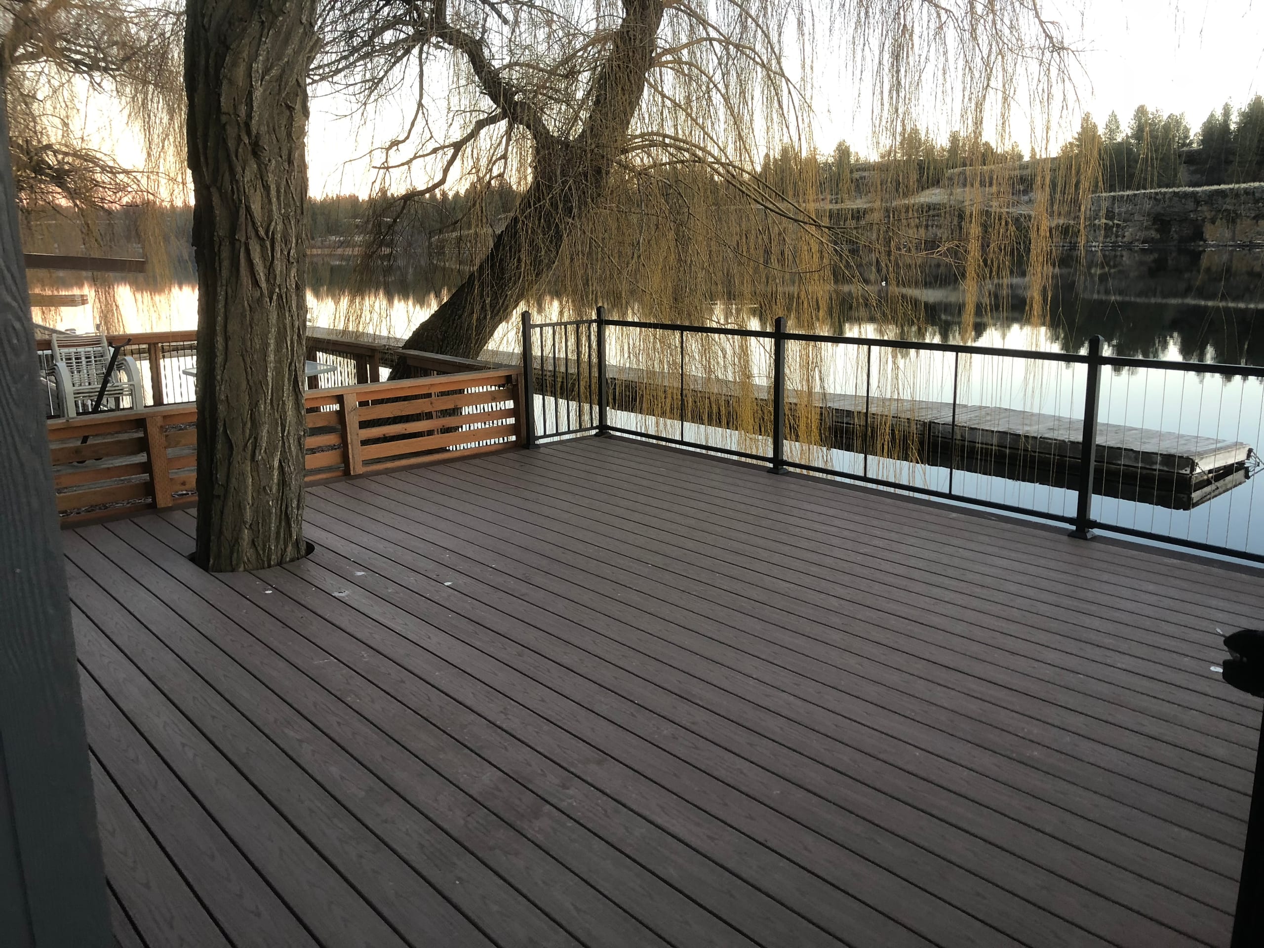 Brown waterfront deck built around a large willow tree, with black metal railing